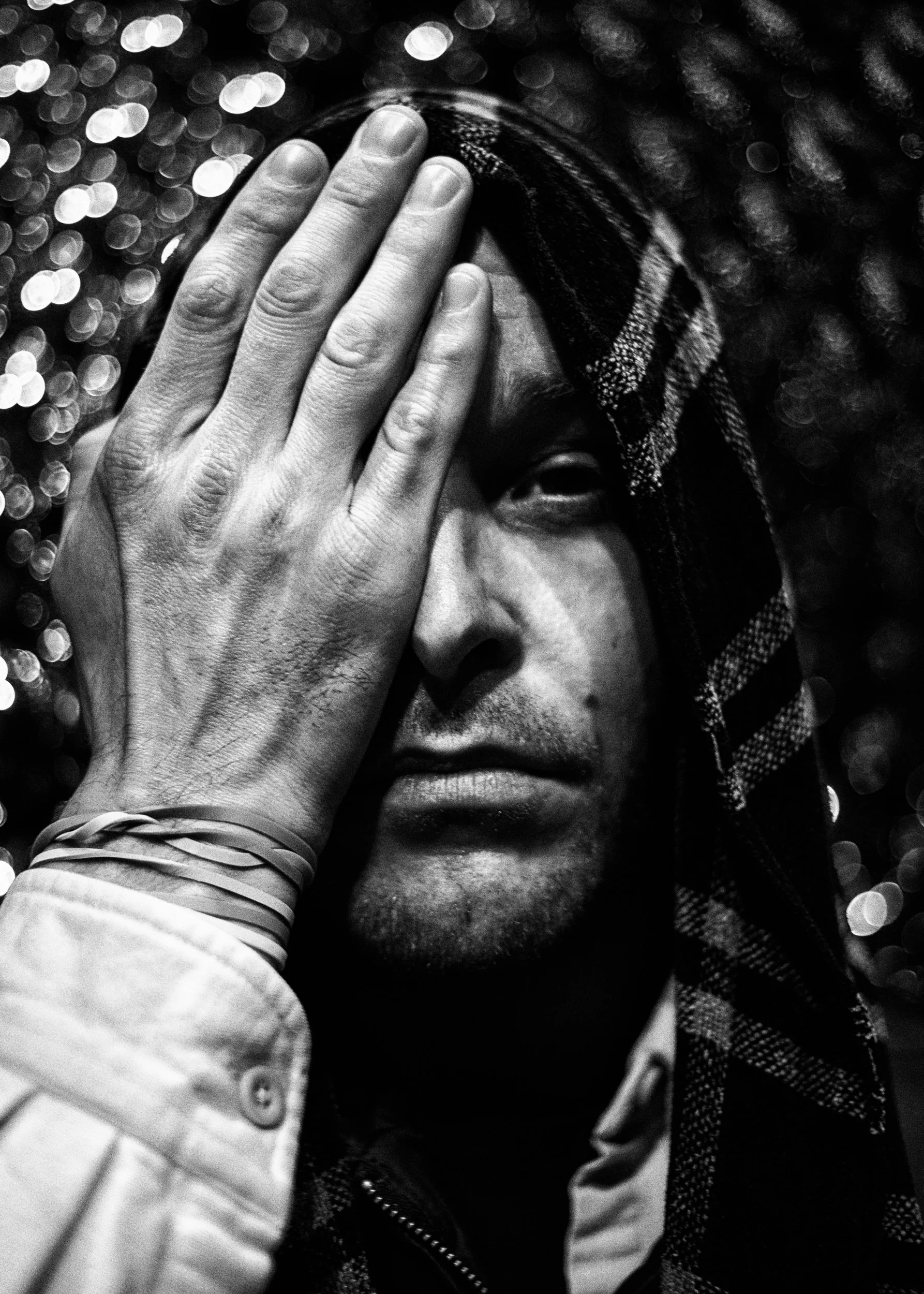 Black and white portrait of a person covering one eye with a hand, wearing a plaid hood, with a bokeh effect background. Daniel Burton, Multi-Disciplinary Artist and Curator