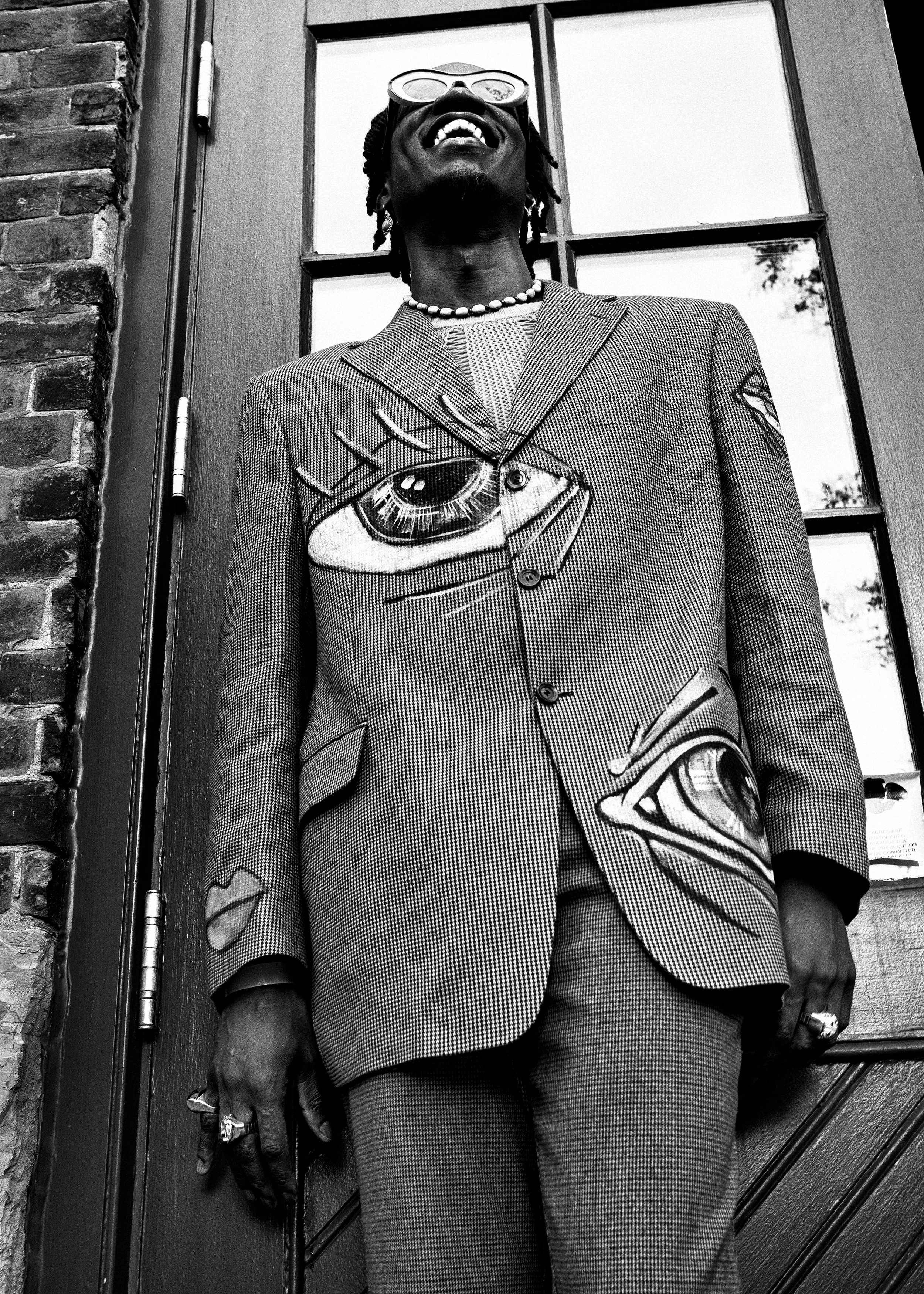 A person wearing a patterned suit with eye-themed embroidery, standing in front of a window and brick wall, laughing and wearing large glasses and jewelry.