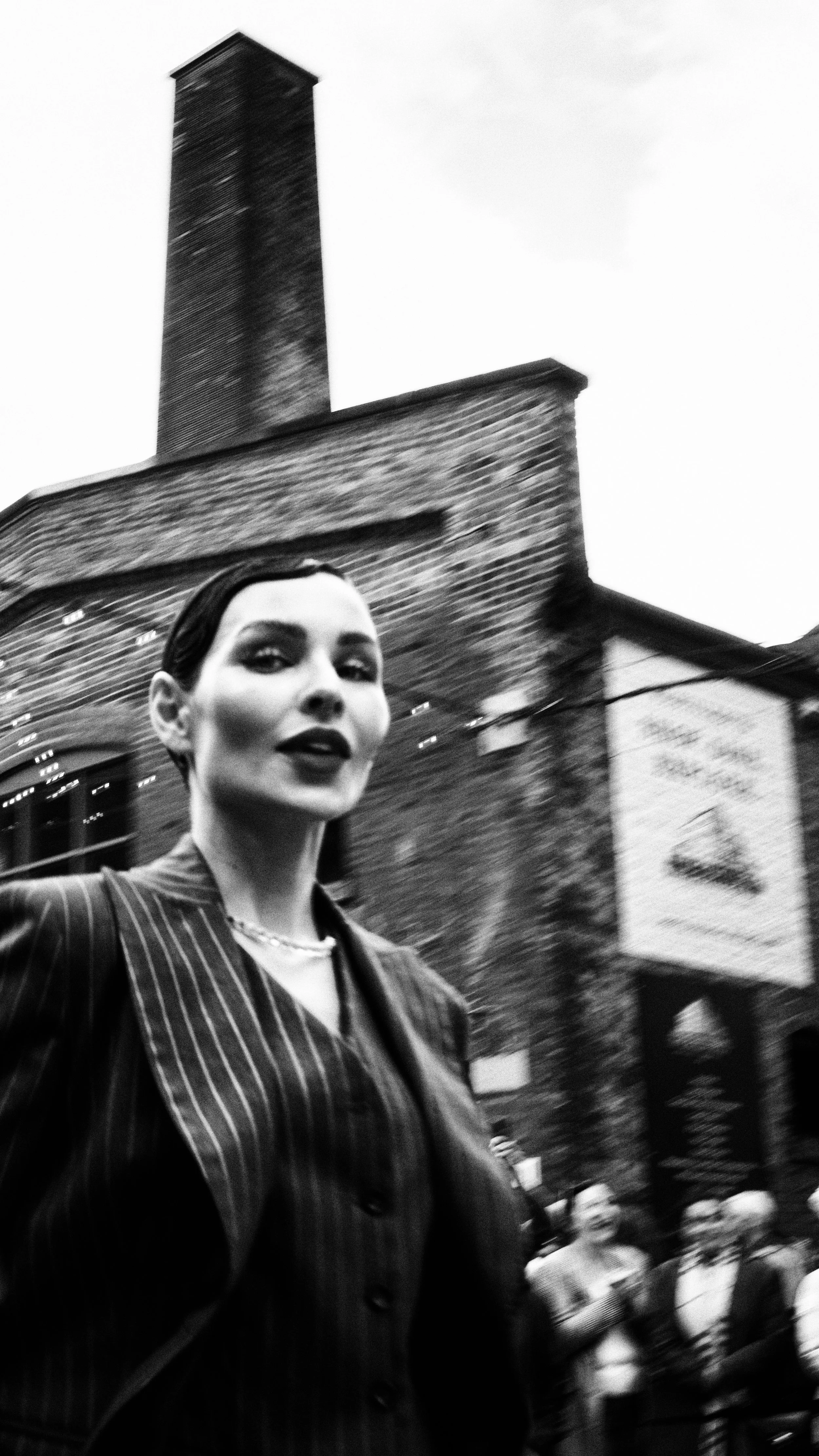 Black and white photo of a woman in a pinstripe suit with slicked-back hair in an urban street scene, with a brick building, chimney, and some out-of-focus people in the background.