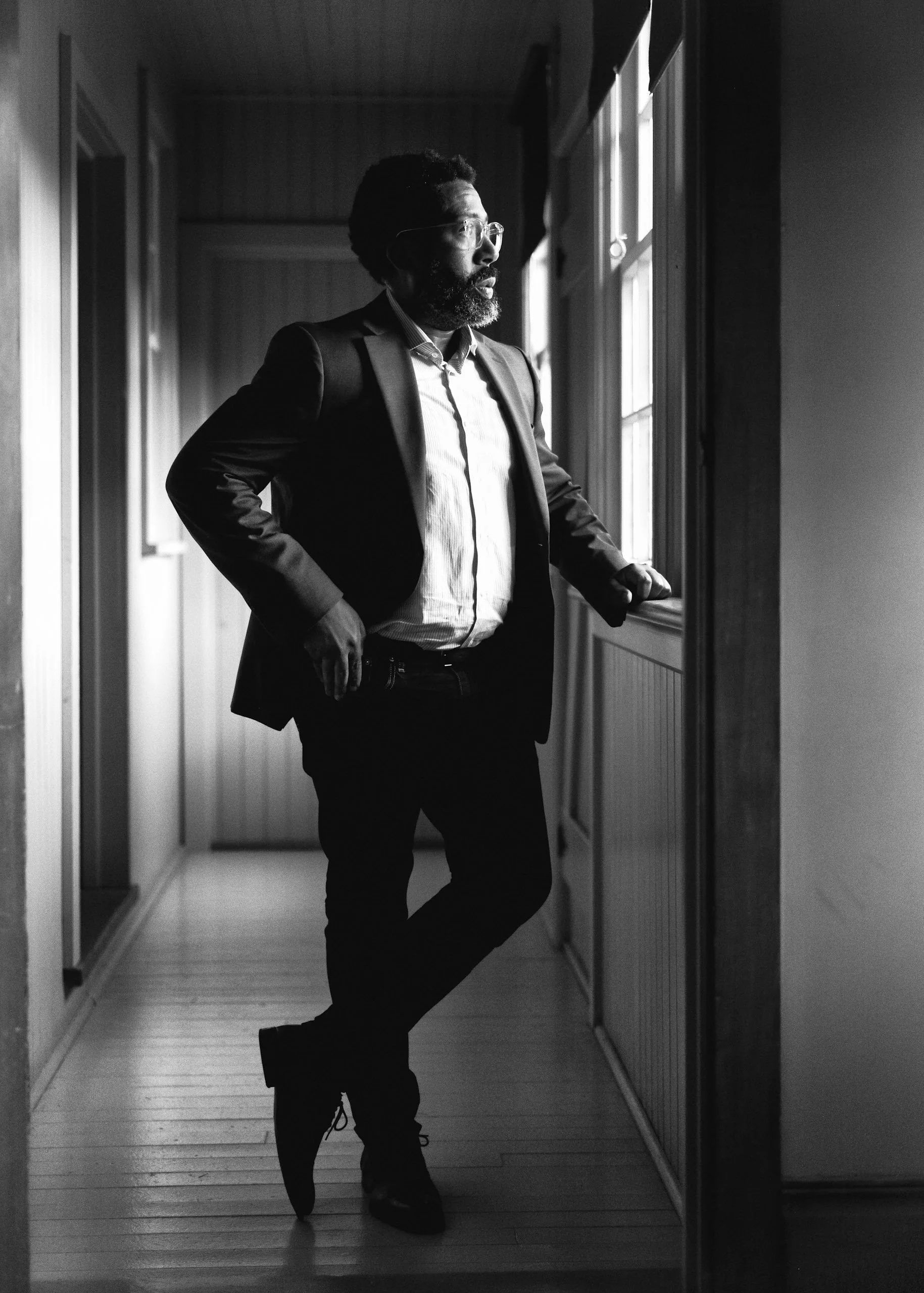 A man with glasses, a beard, wearing a suit jacket and dress shirt, stands in a hallway looking out a window, with one hand on the windowsill and the other on his hip, in black and white.