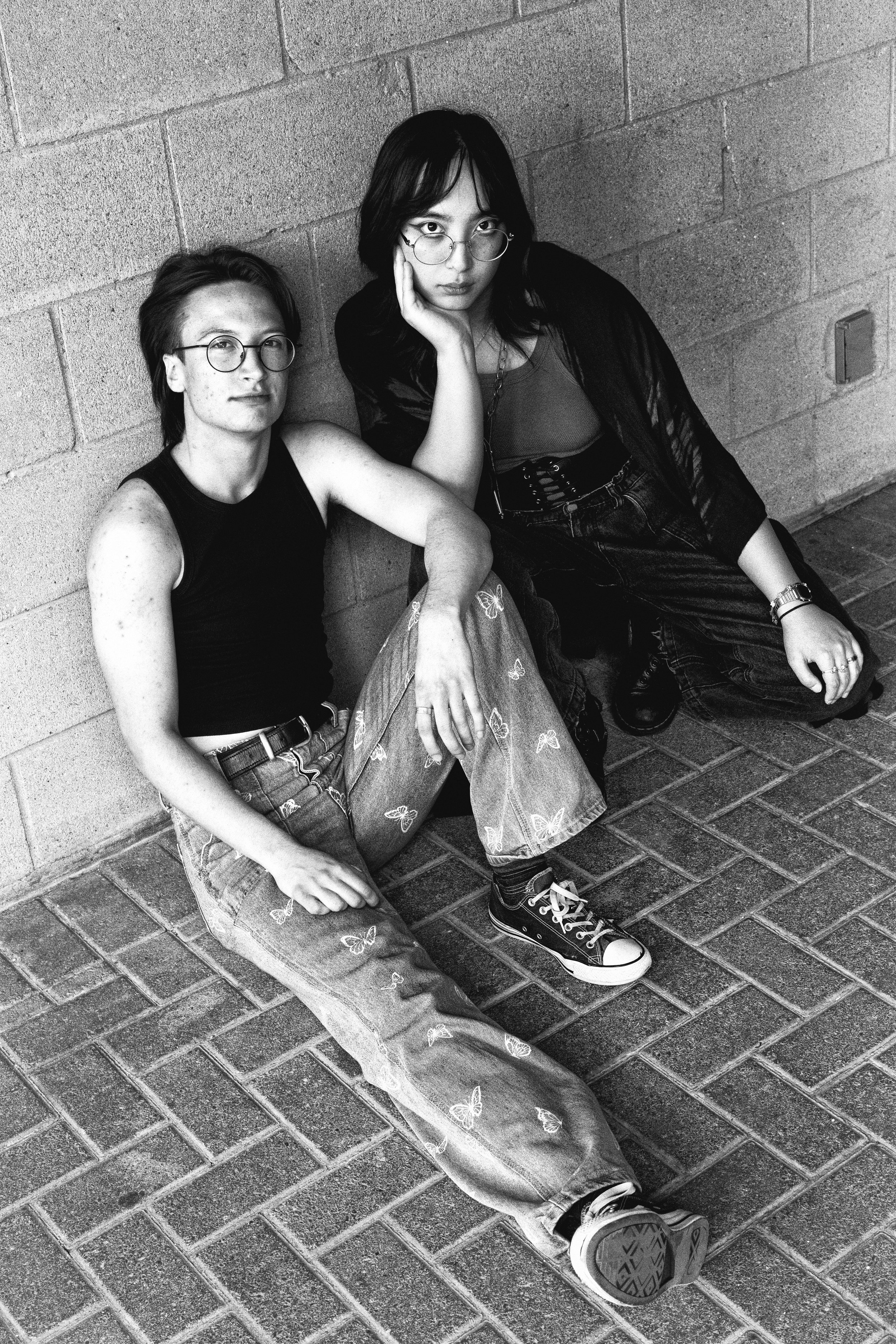Two young women sitting on the sidewalk against a brick wall, both wearing glasses and casual clothing, with one resting her chin on her hand and the other looking directly at the camera.