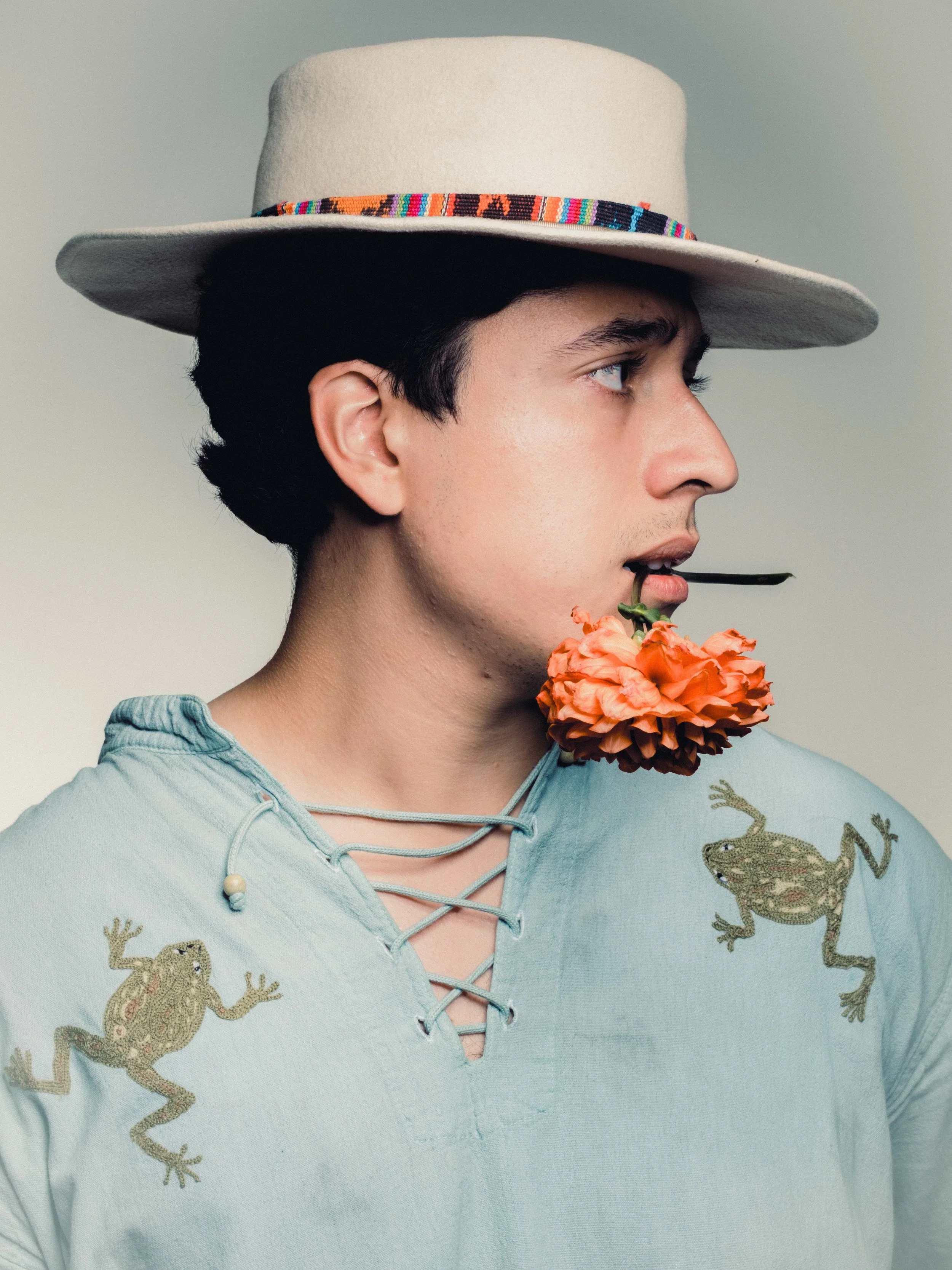 A man wearing a light blue shirt with frog embroidery, a beige hat with a colorful band, holding a flower in his mouth, looking to the side.