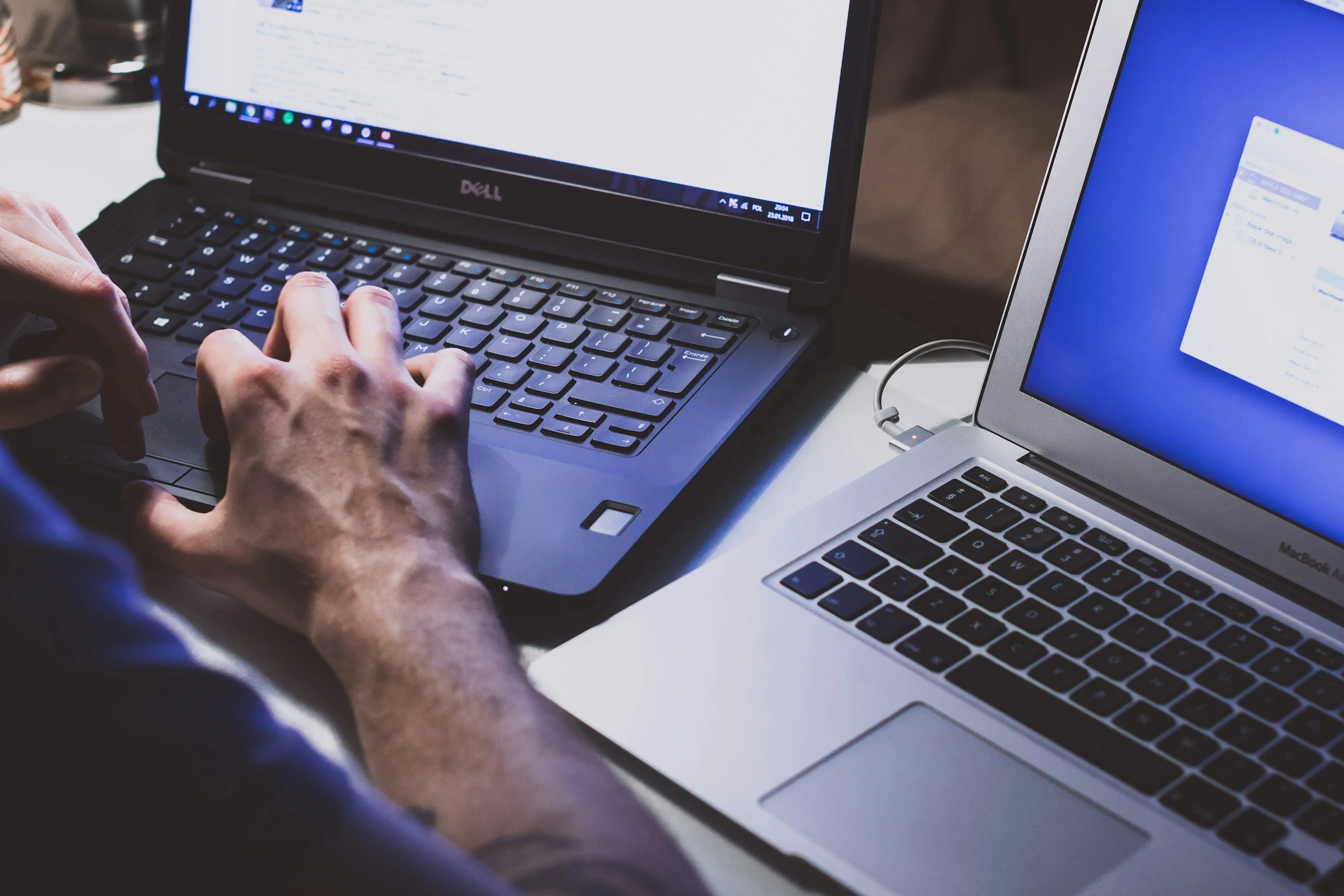 Close-up of hands typing on a black laptop keyboard, with a second laptop displaying a blue screen in the background.