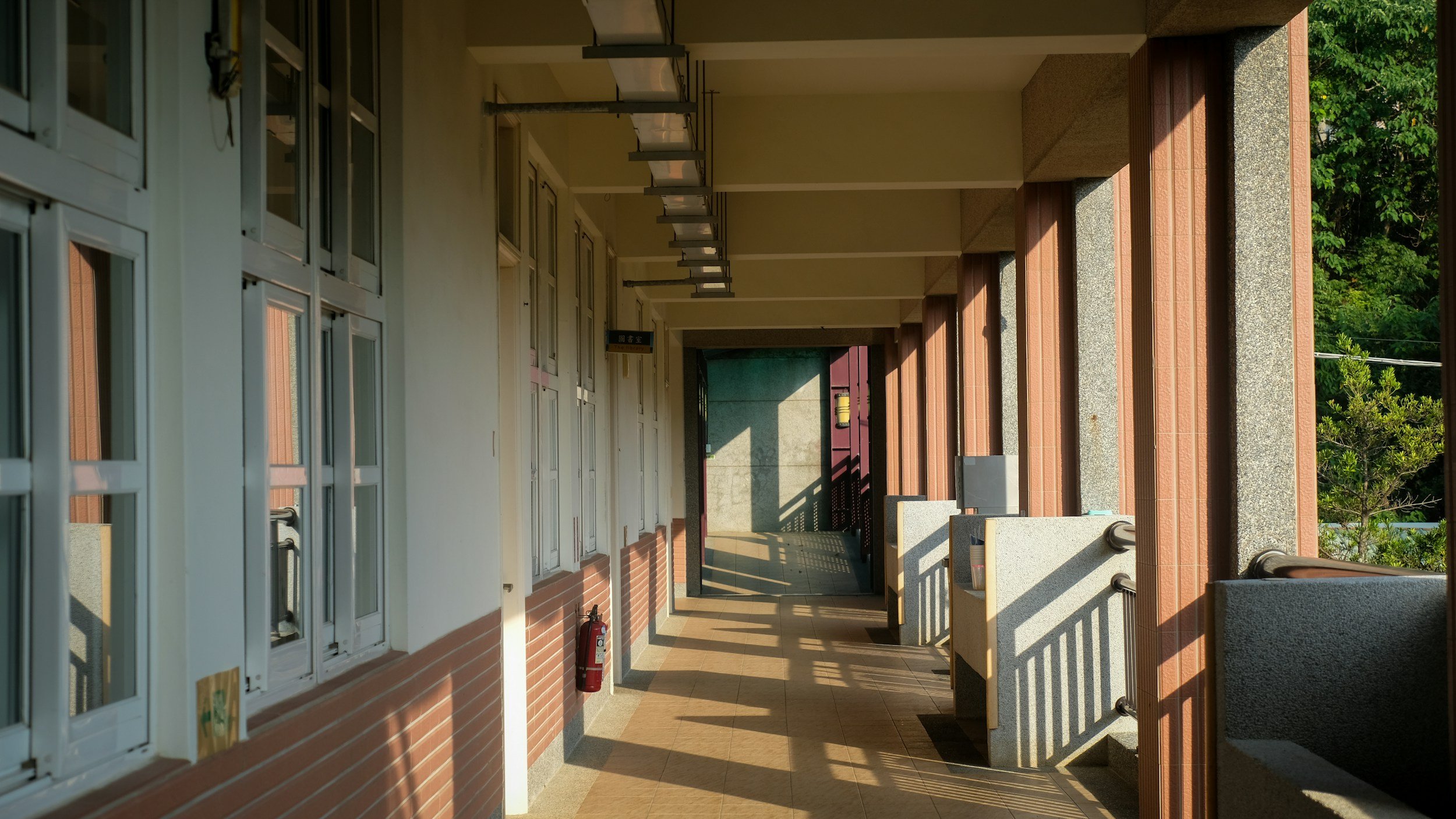 Long outdoor corridor with white windowed doors on the left, brick and concrete walls on the right, a fire extinguisher at the end, and sunlight casting shadows on the floor, greenery visible in the background.
