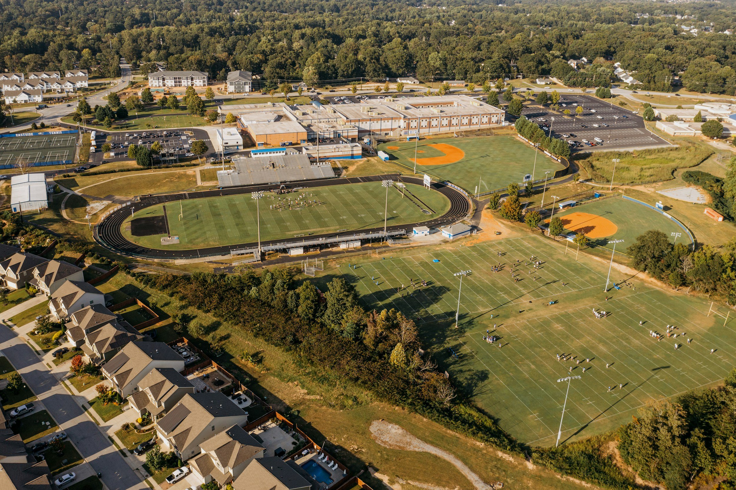 Aerial view of a school campus with sports fields, parking lots, residential houses, and wooded areas in the background.