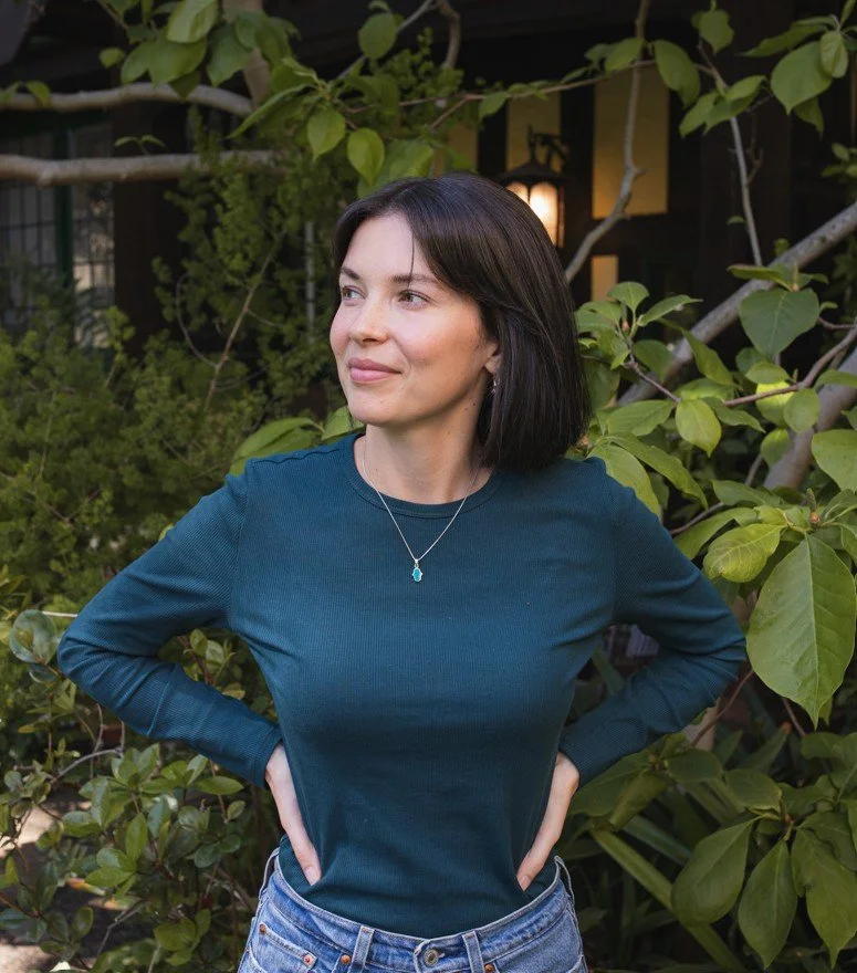 Woman with short dark hair wearing a teal long-sleeve shirt and jeans, standing outdoors among green foliage.