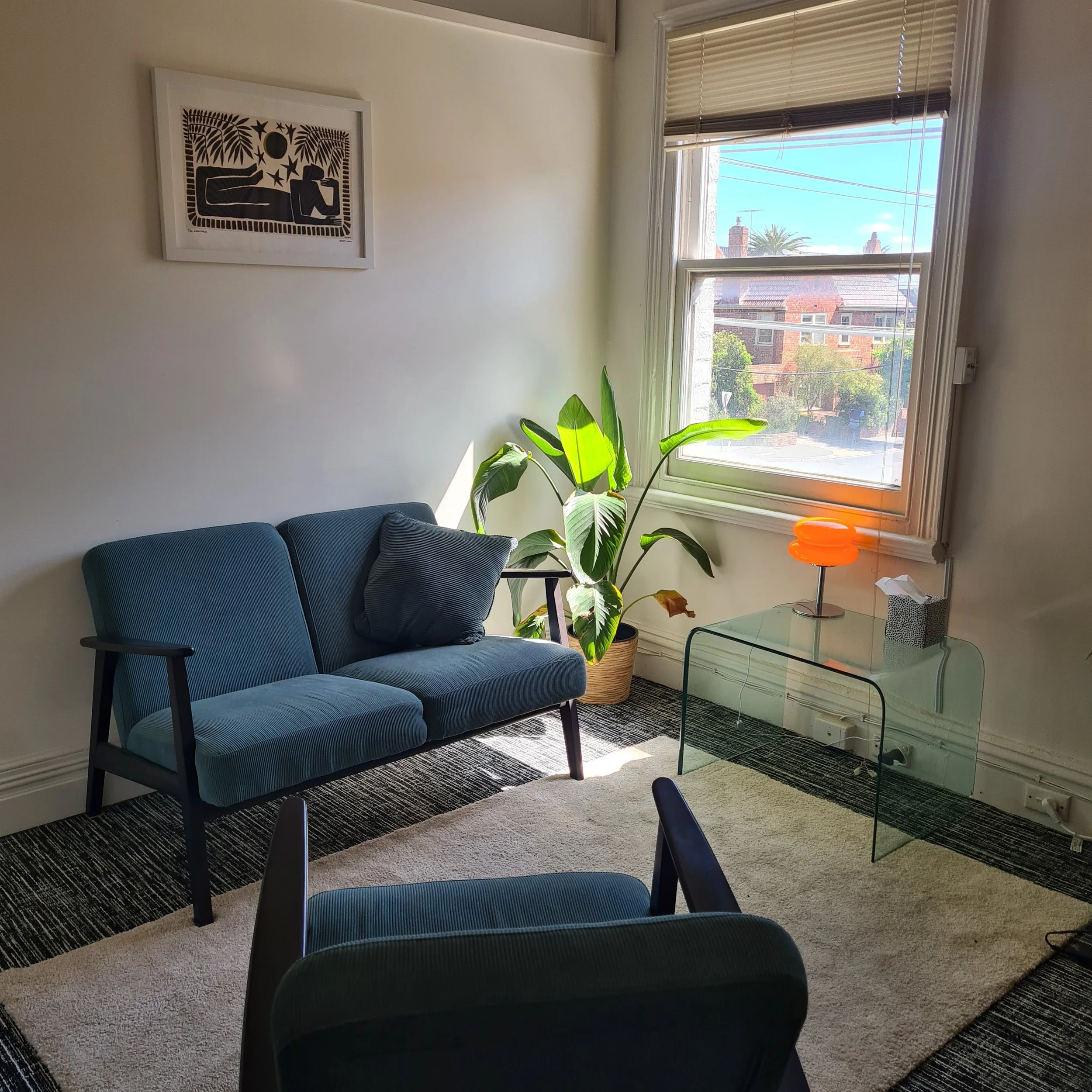 A cozy living room corner with a blue sofa, a black cushion, a leafy green potted plant near a window, a glass side table with an orange lamp, a small tissue box, a framed artwork on the wall, and a mix of beige and dark patterned rugs.