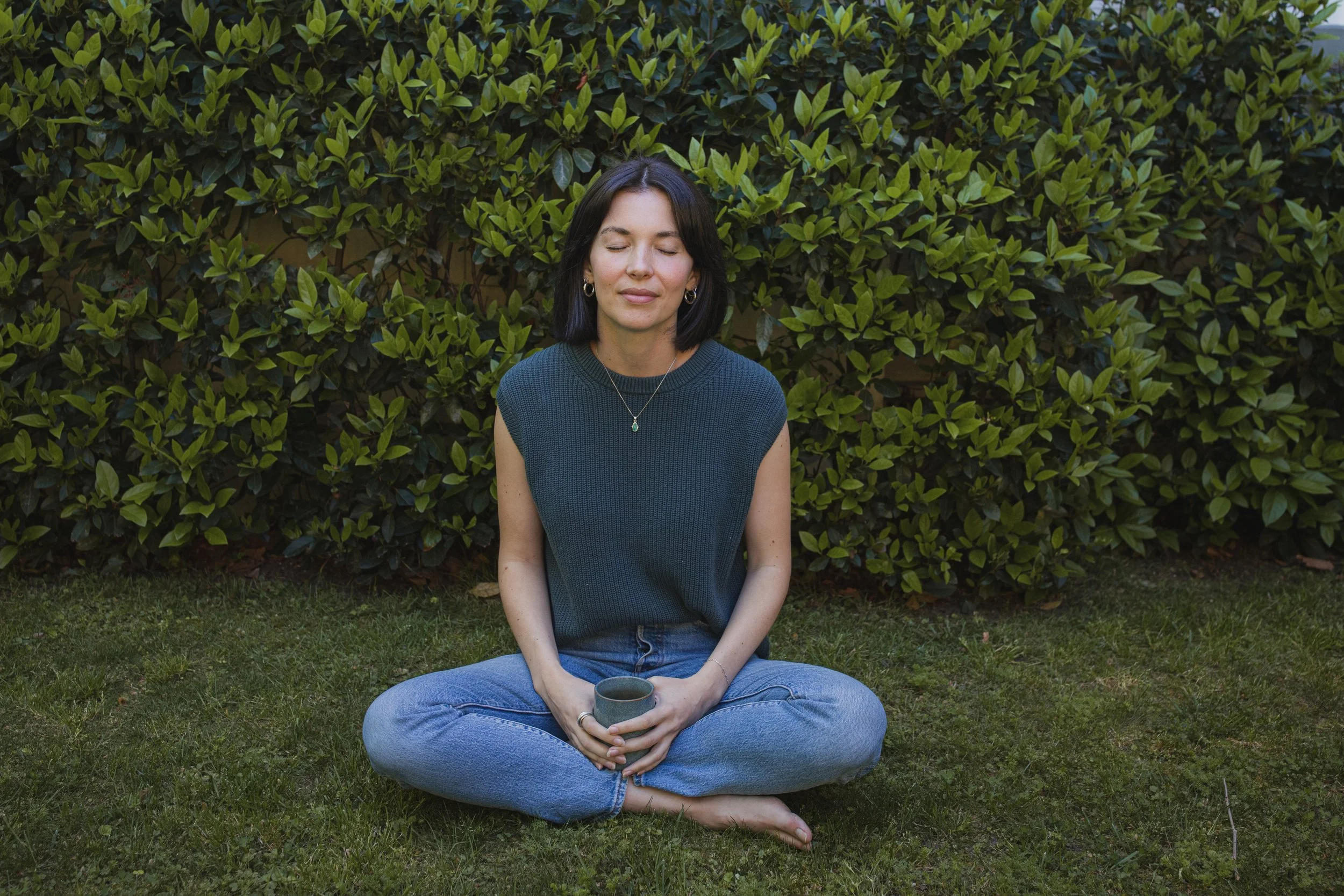 A woman sitting cross-legged on the grass outdoors, holding a cup with her eyes closed, with green bushes behind her.