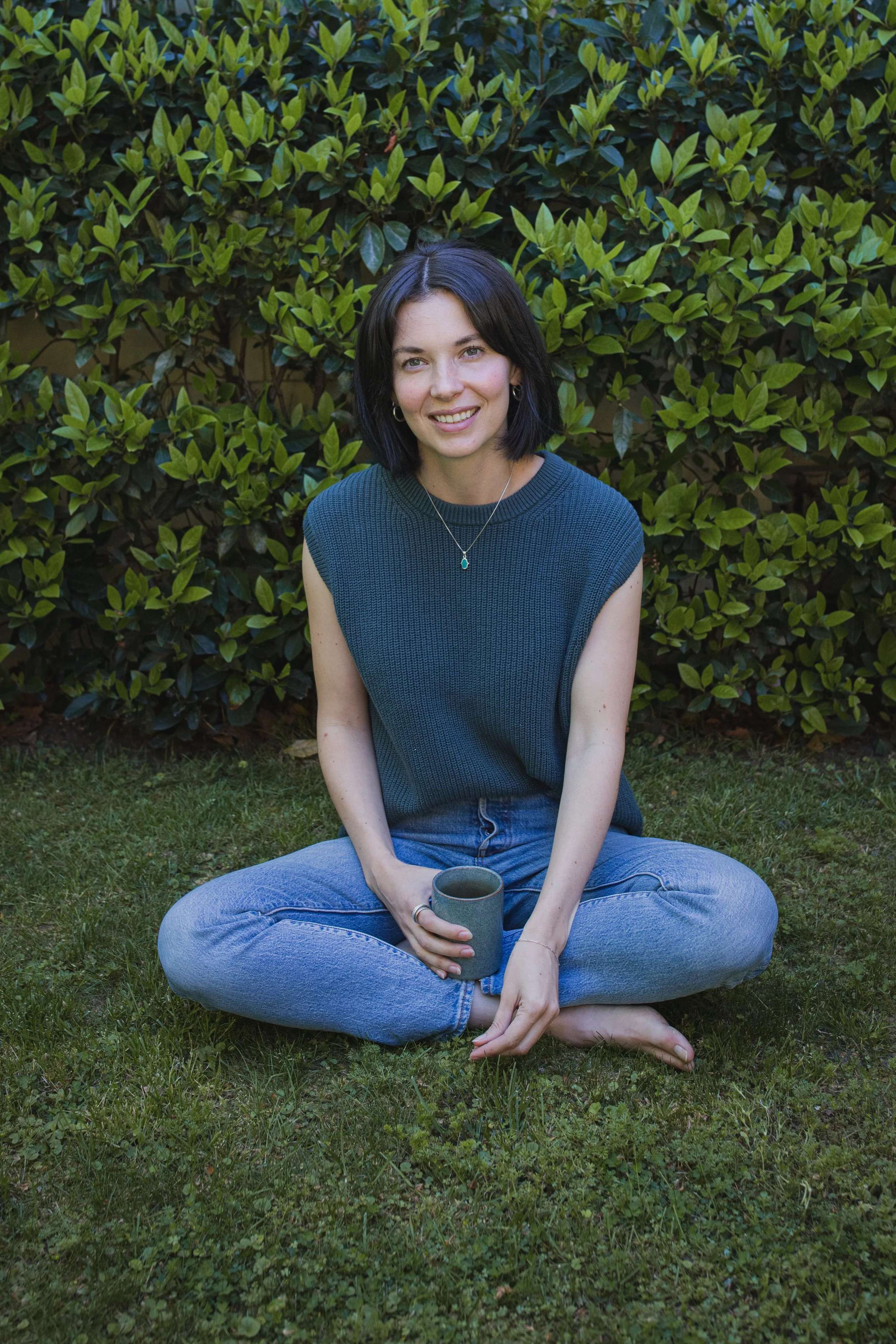 A young woman with shoulder-length dark hair, sitting cross-legged on grass in front of a leafy green bush, smiling at the camera, holding a gray mug.