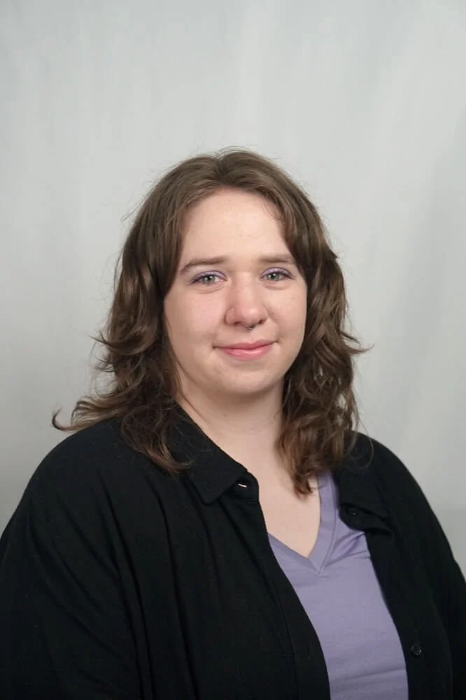 A young woman with shoulder-length curly brown hair, wearing a black shirt and a lavender top, standing against a plain light gray background.