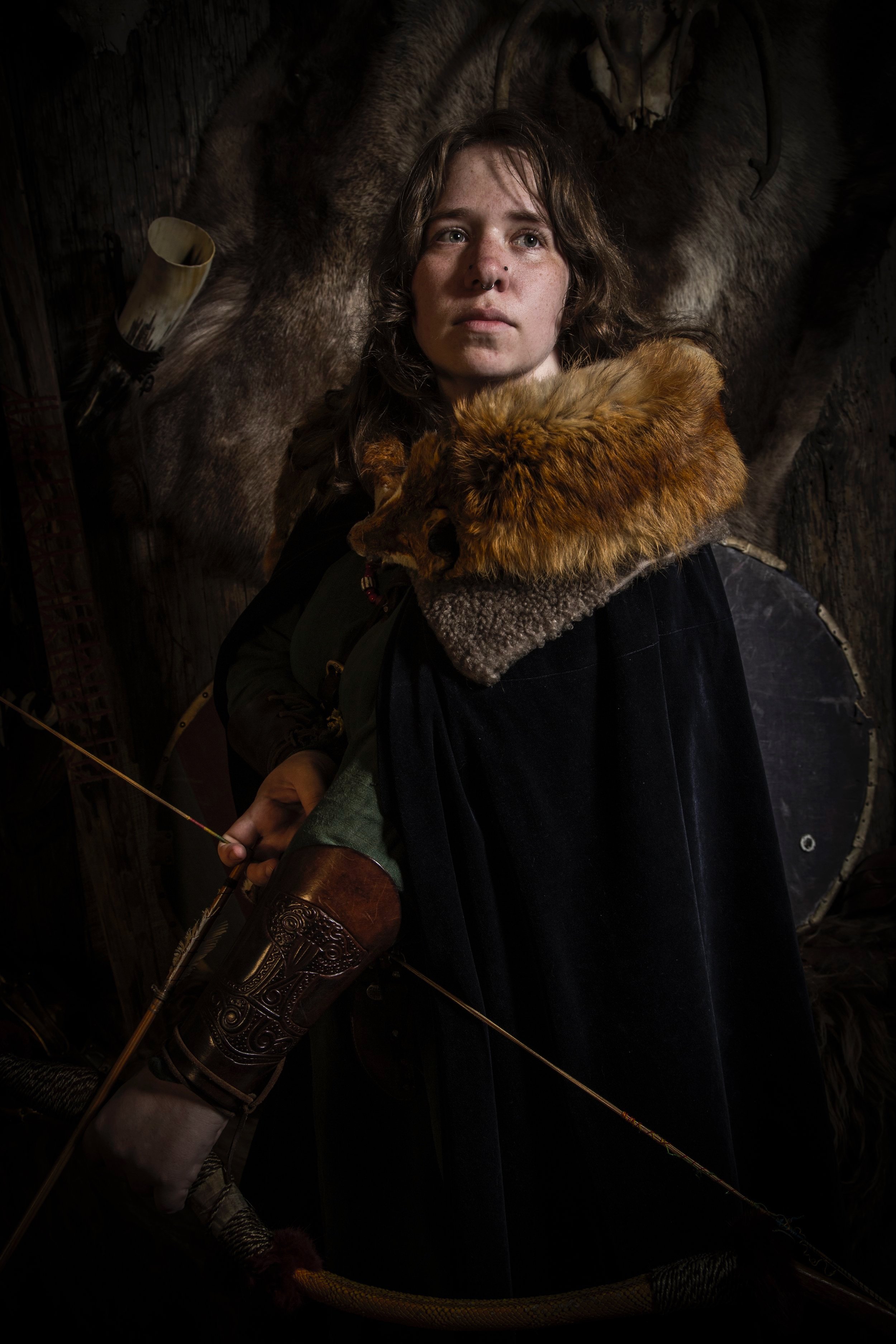 A person in medieval/fantasy attire, with a fur-lined cloak, holding a bow, standing in front of a mounted animal hide background.