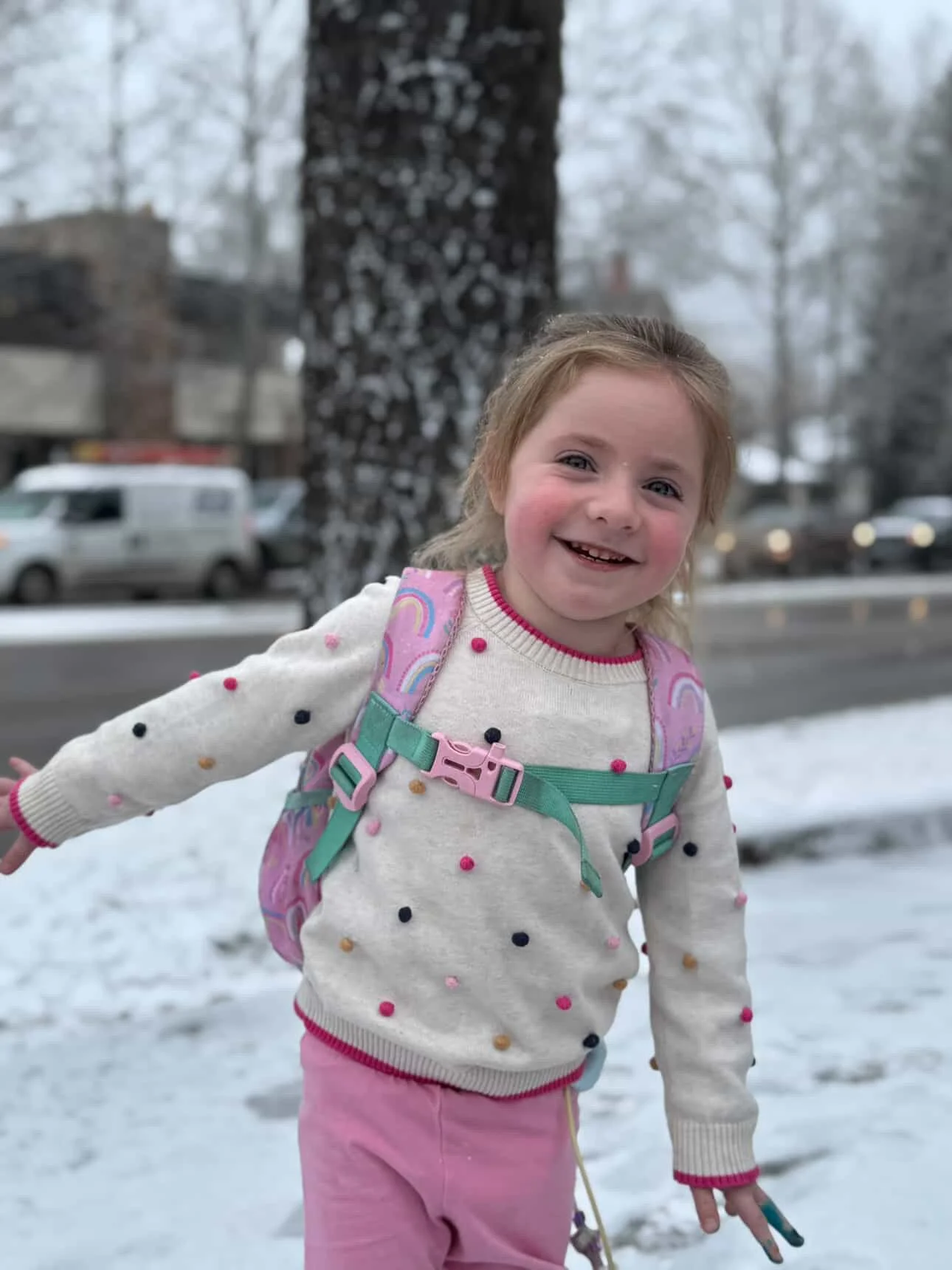 A young girl with rosy cheeks and a button feeding tube smiling outside in the snow, wearing a white pom-pom sweater, pink pants, and a pink rainbow backpack.