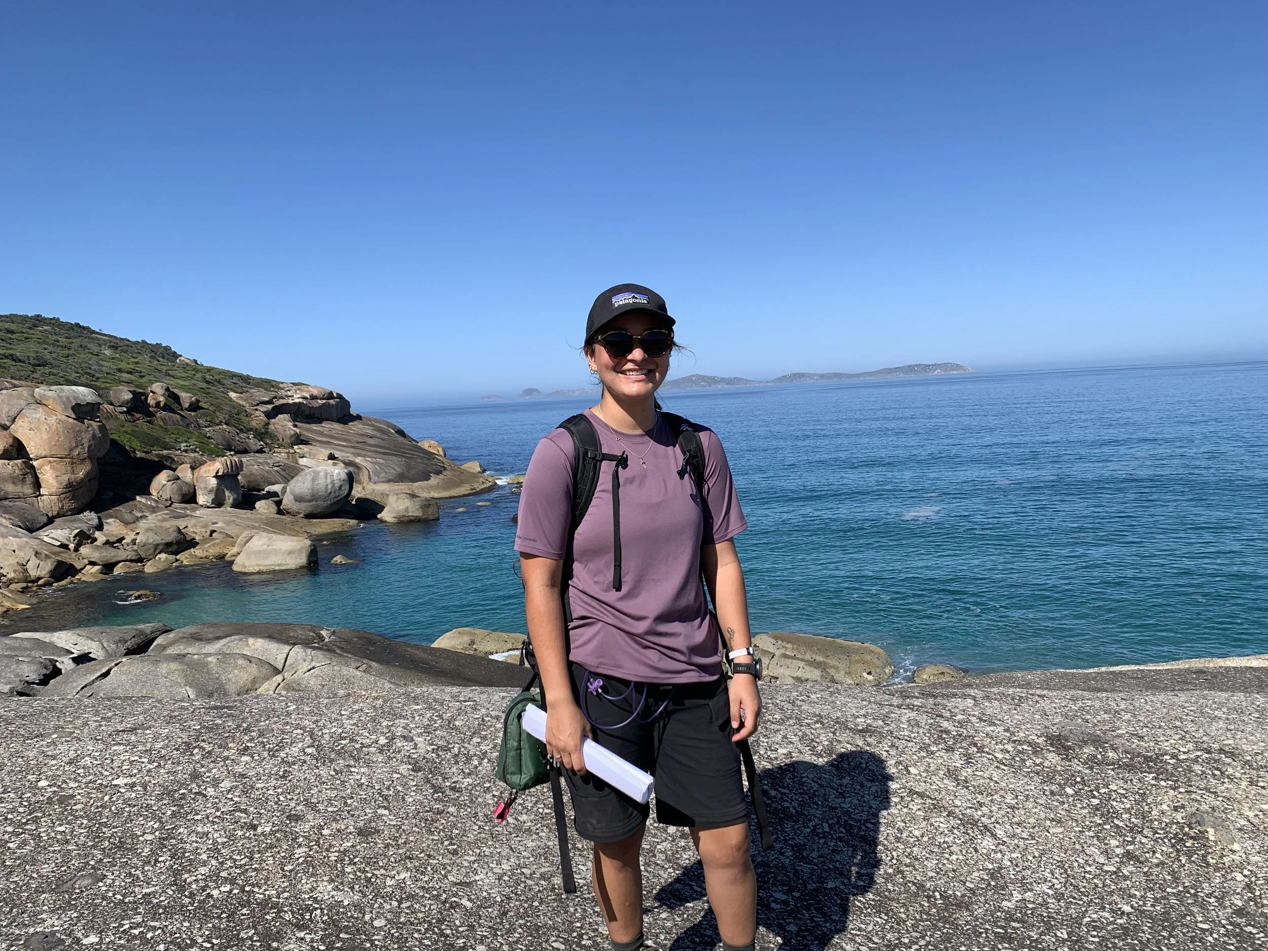 A woman in a cap and sunglasses smiles while hiking, standing on granite rocks with a backpack and hydration pack, overlooking a turquoise coastal bay with distant hills on the horizon.