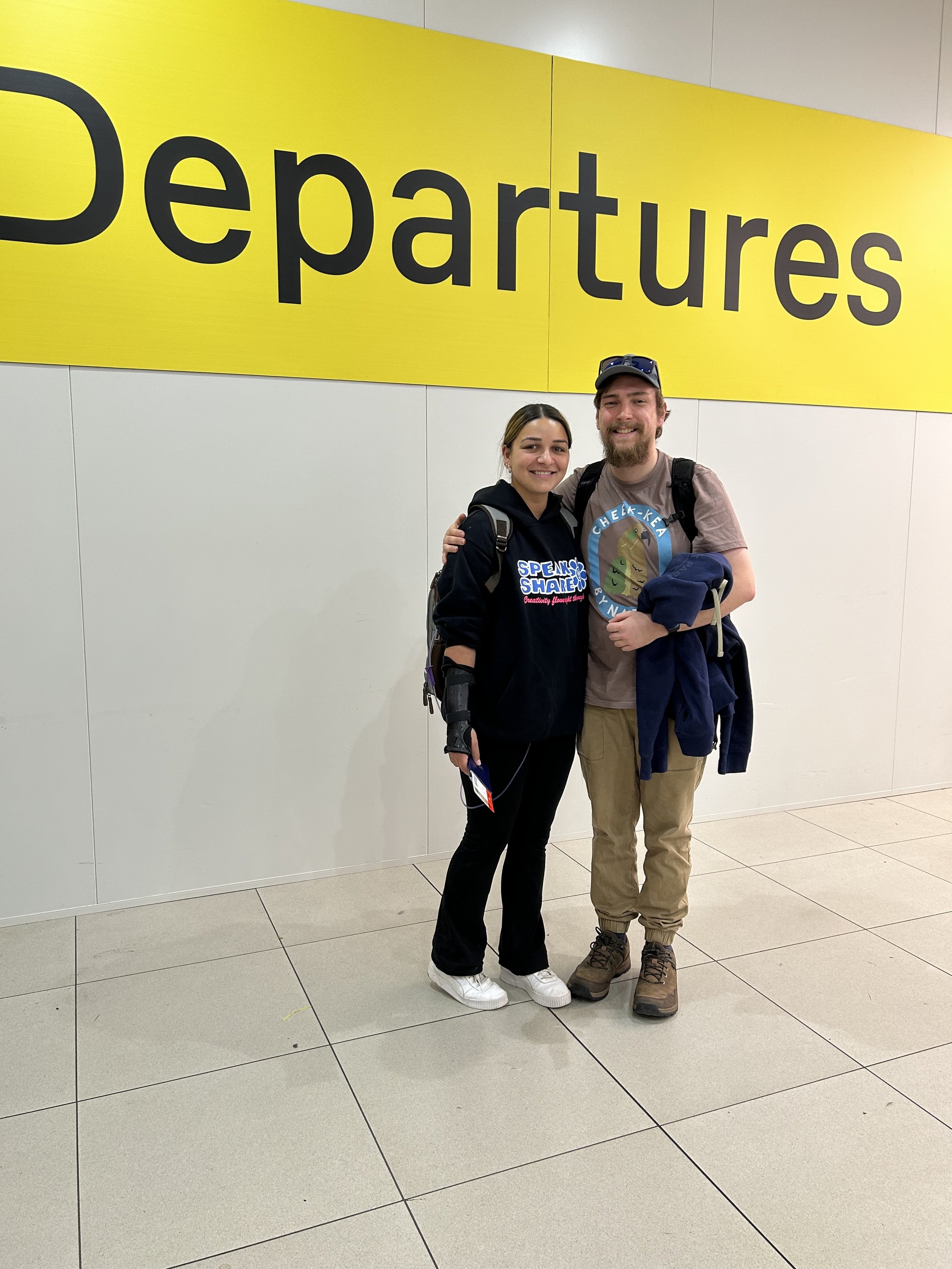 A woman and man smile with arms around each other in an airport, standing below a large yellow "Departures" sign.