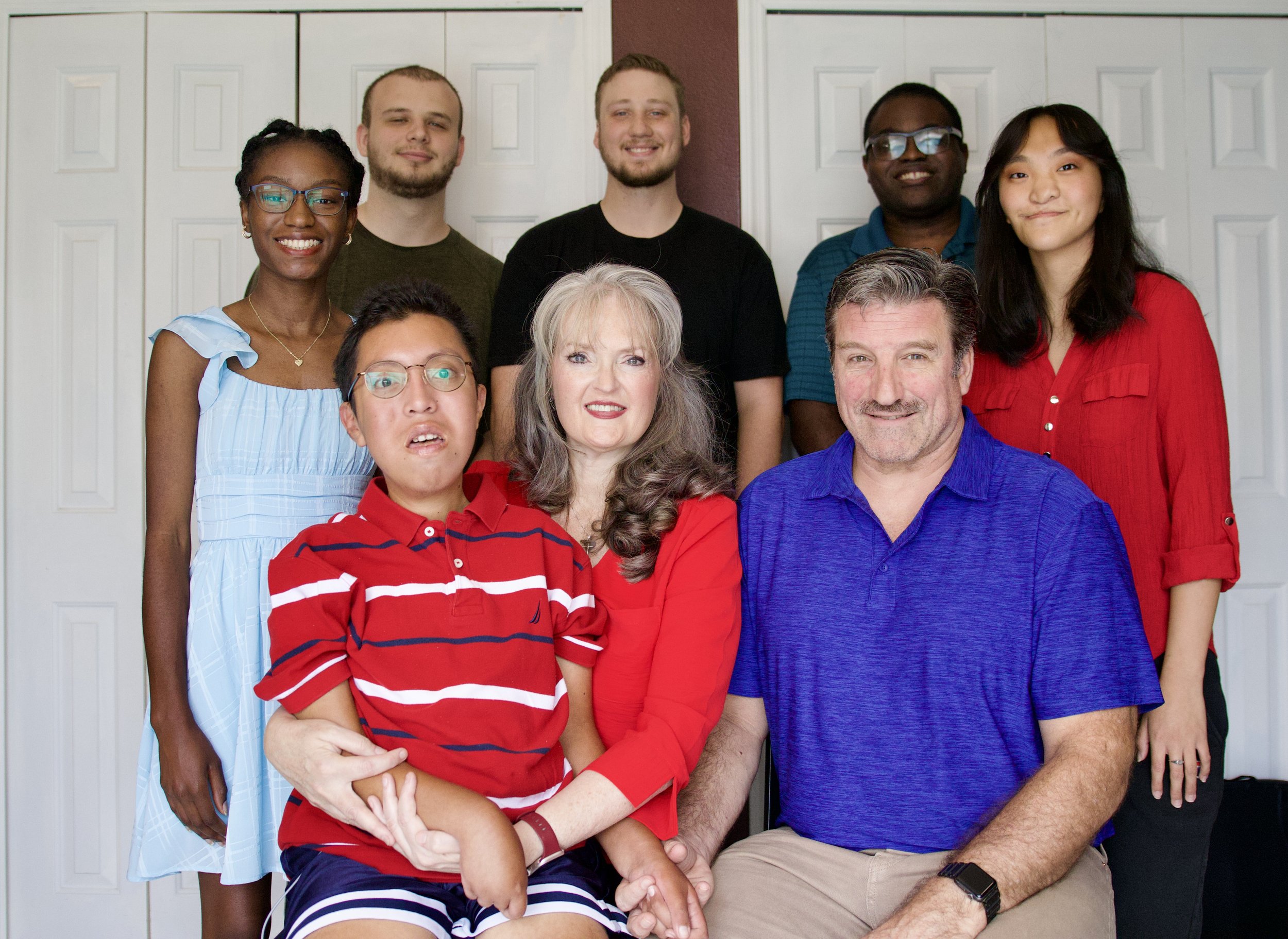 A family of eight — two adults and six young adult and teenage children of different ethnicities — pose together smiling in front of white doors.