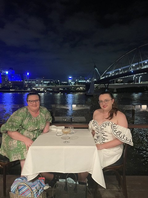 Two women sitting at a table by the waterfront at night, with an illuminated bridge in the background, one wearing a green patterned dress and the other in a white outfit with a nasogastric tube, both wearing glasses and smiling at the camera.