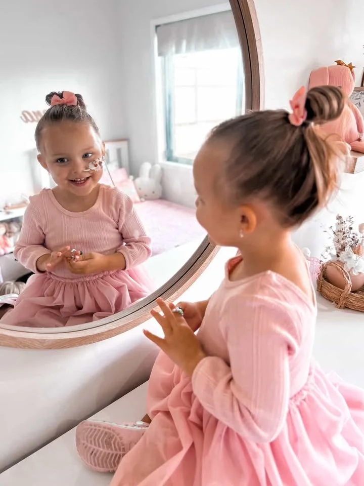A young girl in a pink dress with a bow in her hair looking into a mirror, smiling at her reflection.