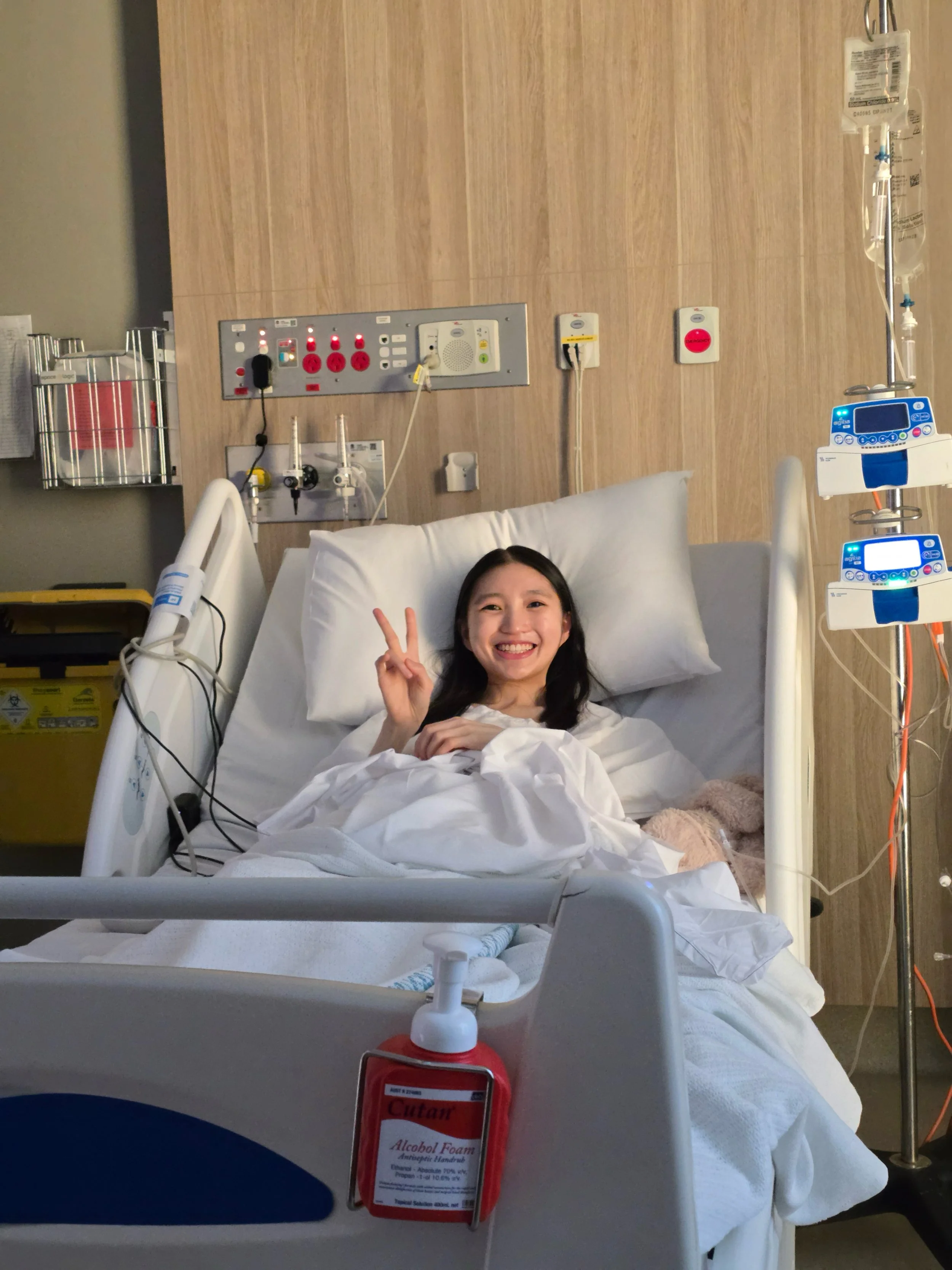 Zi Ting lies in a hospital bed giving a peace sign to the camera, smiling broadly, with medical equipment and IV lines visible beside her.