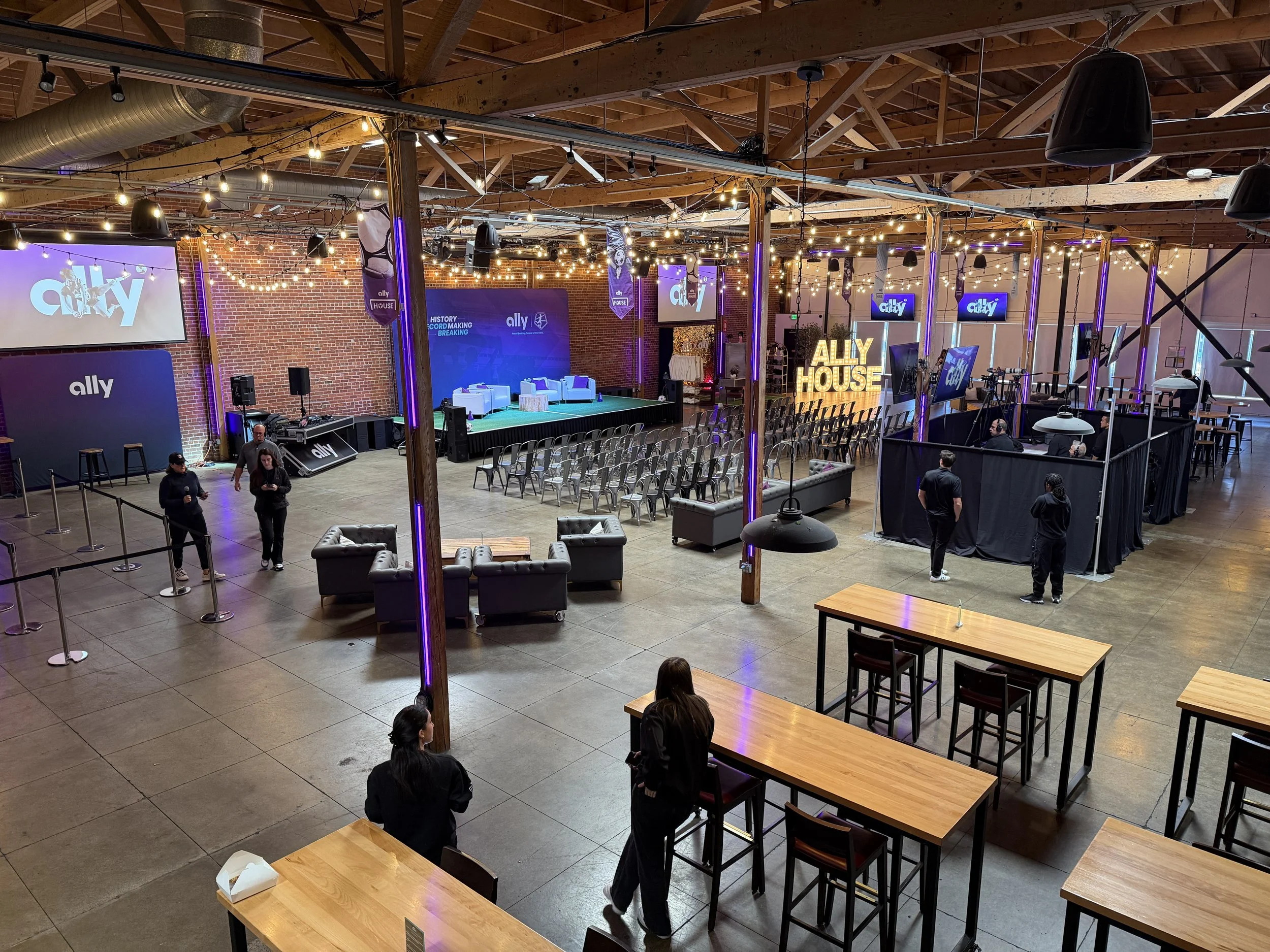 An event space with a stage, multiple screens displaying the word 'ally', and seating arrangements. The room has industrial-style decor with brick walls and exposed wooden beams, decorated with string lights.