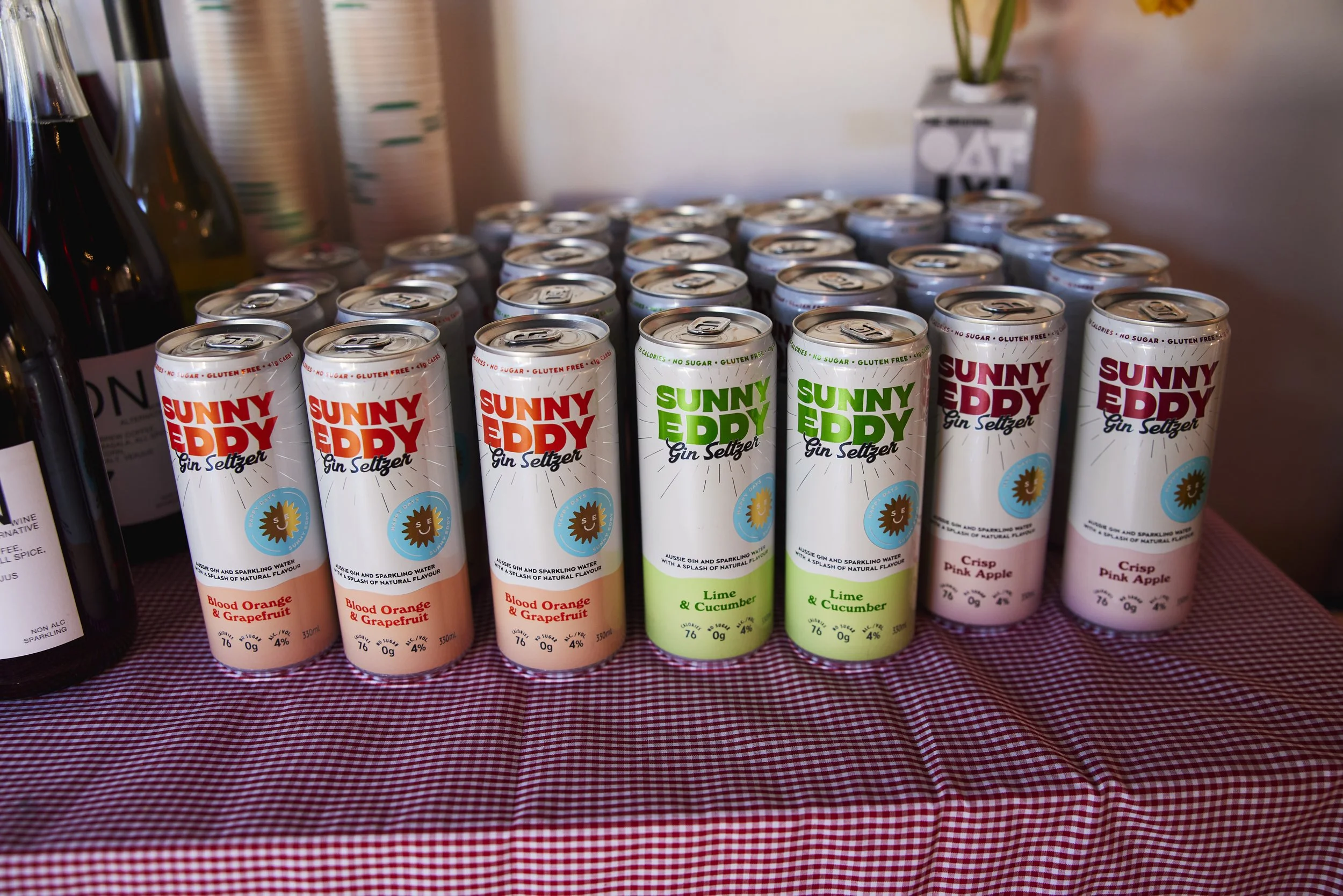 Cans of Sunny Eddy Gin Seltzer in flavors Blood Orange & Grapefruit, Lime & Cucumber, and Crisp Pink Apple on a table with a checkered cloth.