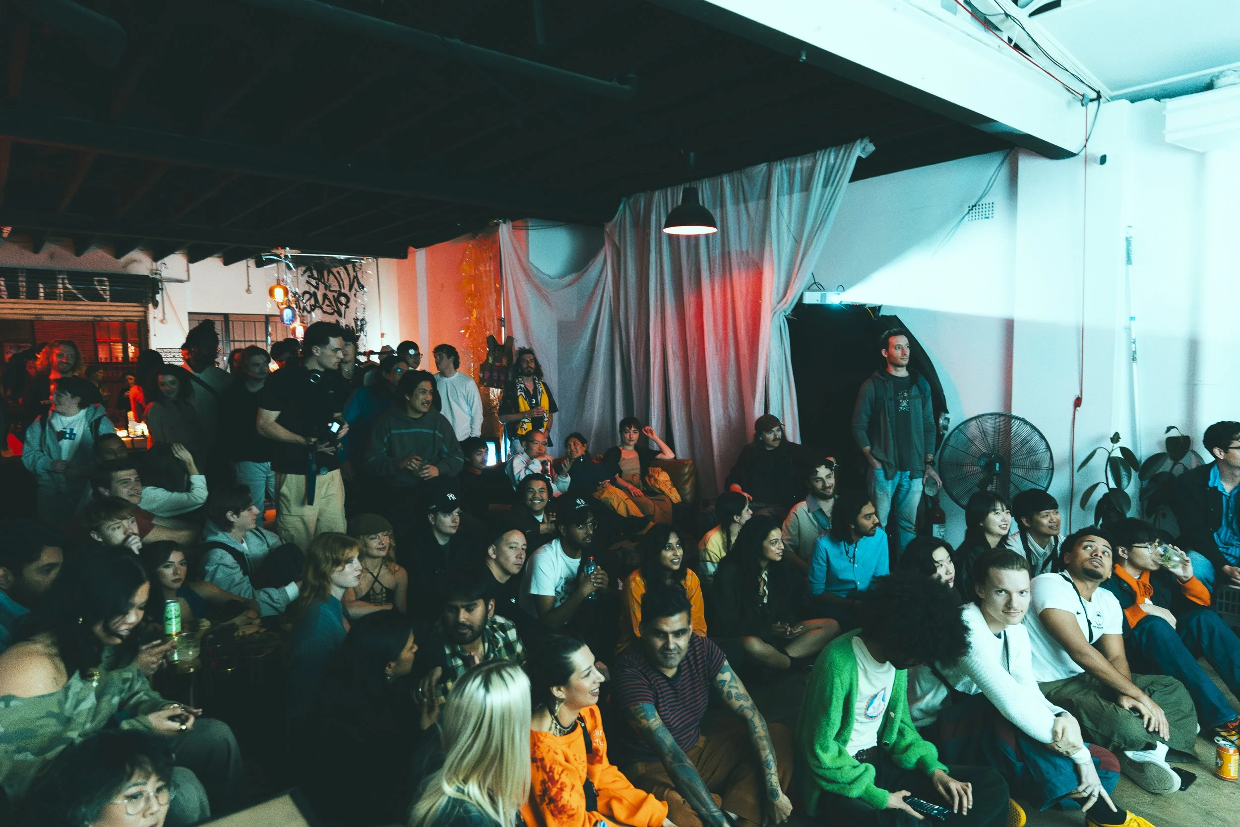 Gathering of people in a dimly lit room with various attendees sitting and standing, including a fan and draped curtains in the background. Attendees are viewing a short film screening.