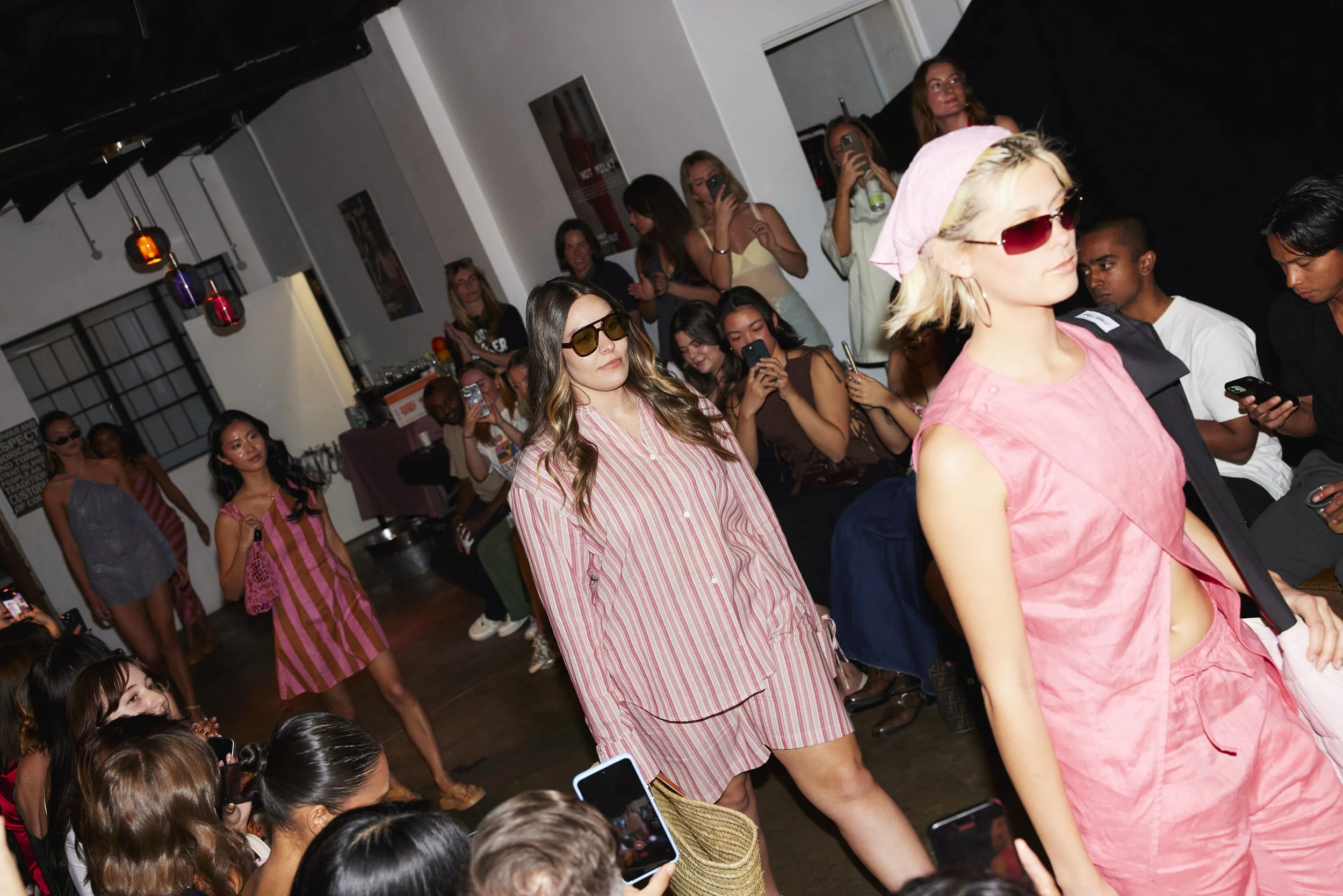 Fashion show with models on runway, audience taking photos, diverse outfits, indoor event.