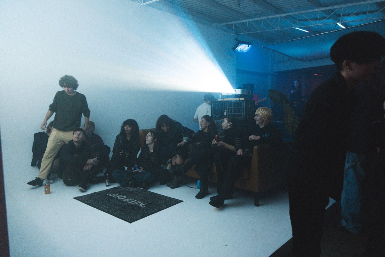 Group of people sitting in a dimly lit indoor space with a projector and a 'Keep Off' rug.
