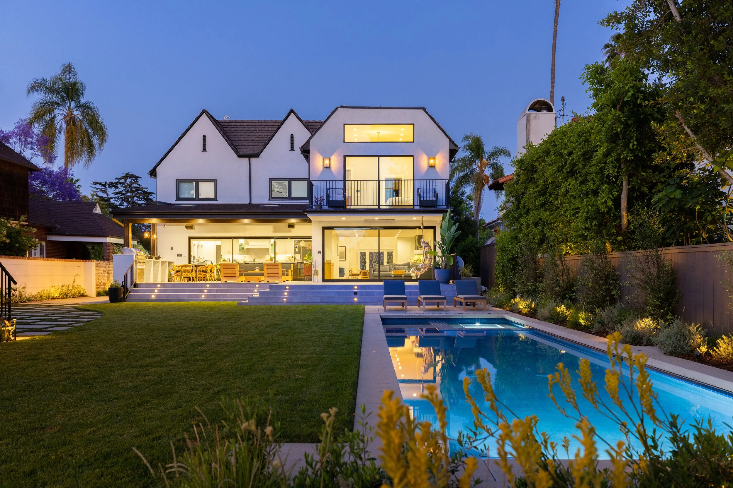 Golf course estate with clean lines against classic Tudor, layered landscaping, linear pool, driveway, lush lawn and warm landscape lighting.