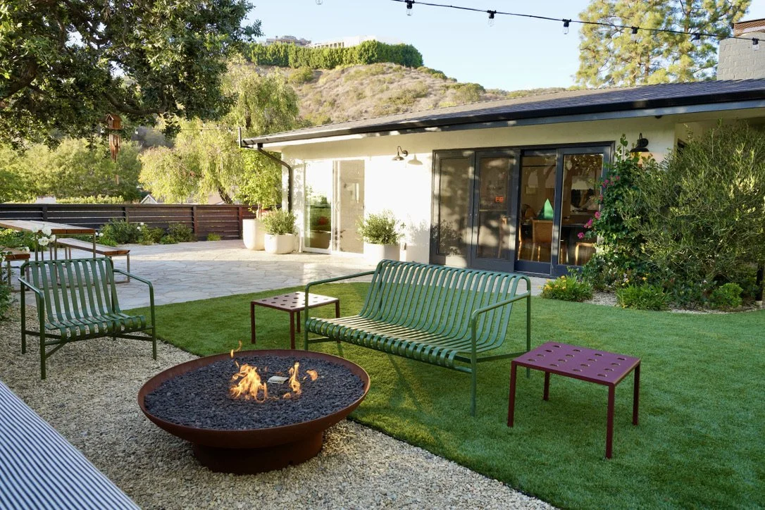 Cozy and bright yard with lush synthetic turf, fire pit lounge, play structure, horizontal slat fencing, decorative potted plantings, layered perimeter landscaping, Mexican pebbles.