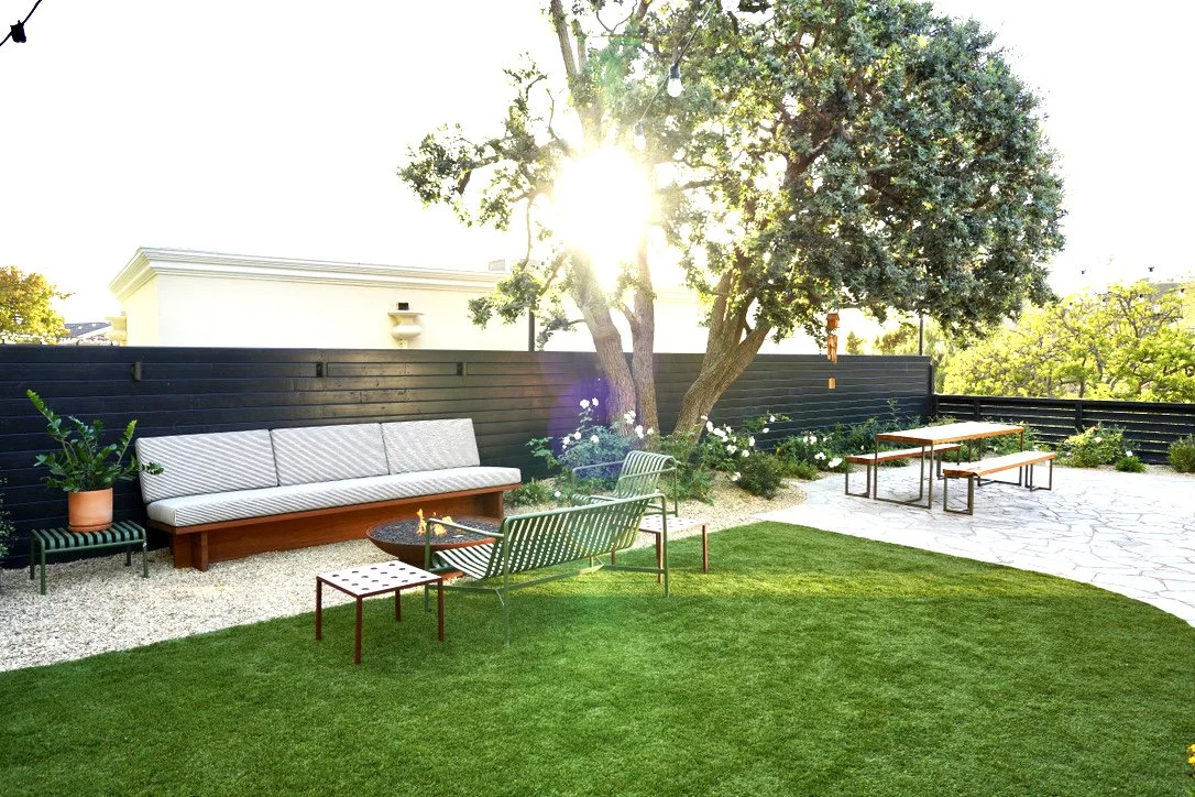 Cozy and bright yard with lush synthetic turf, fire pit lounge, play structure, horizontal slat fencing, decorative potted plantings, layered perimeter landscaping, Mexican pebbles.