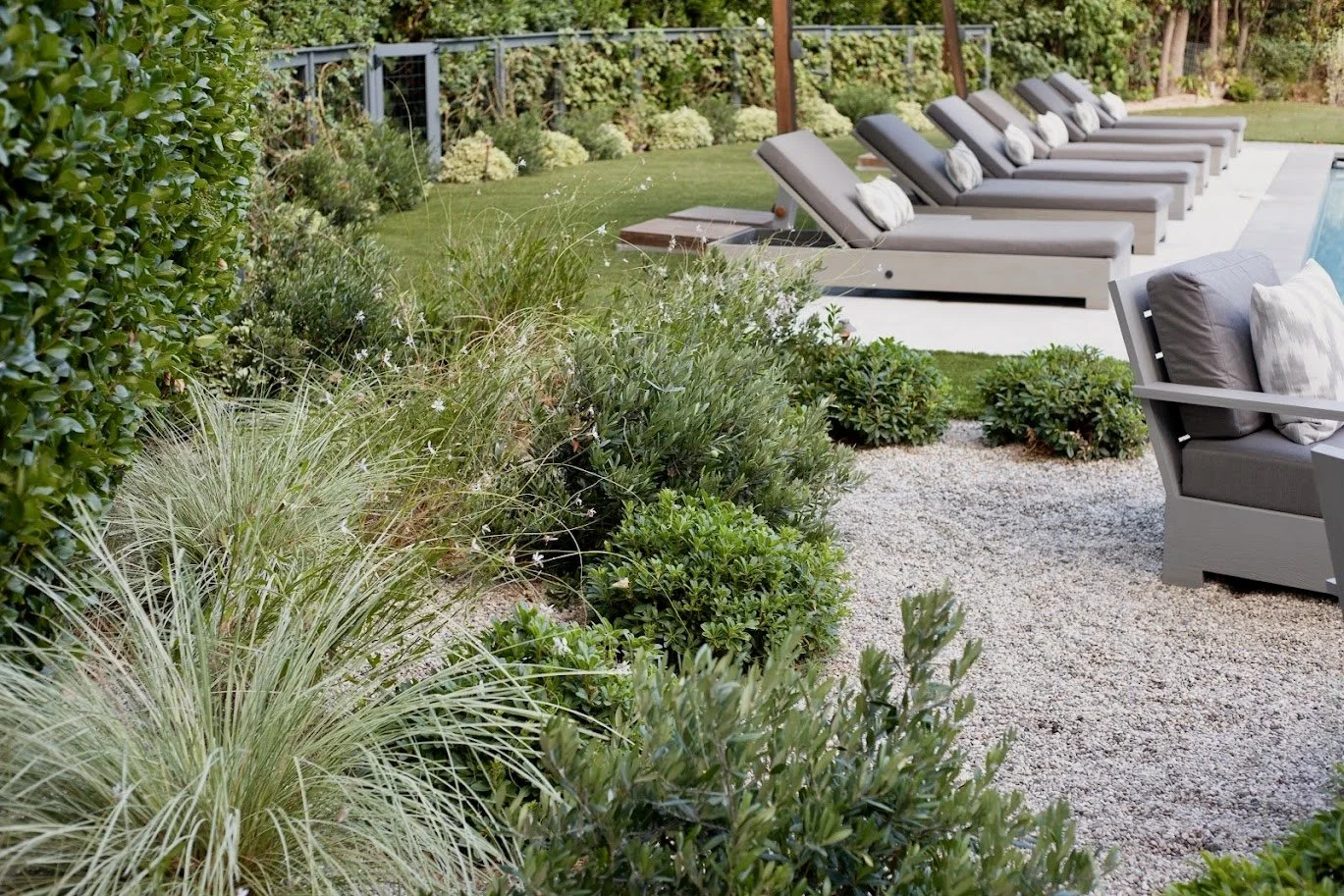 Large entertainer's yard with linear pool, chaise lounges, cantilevered umbrellas, fire pit gravel lounge, lush layered Mediterranean plantings and privacy hedges, front sitting porch, walkways through the garden.
