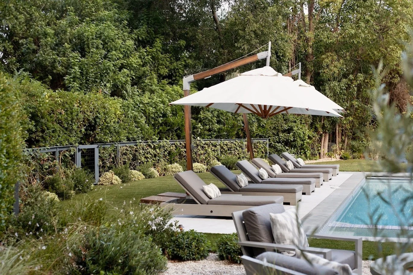 Large entertainer's yard with linear pool, chaise lounges, cantilevered umbrellas, fire pit gravel lounge, lush layered Mediterranean plantings and privacy hedges, front sitting porch, walkways through the garden.