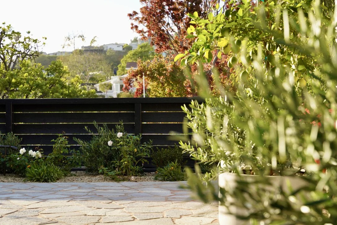 Cozy and bright yard with lush synthetic turf, fire pit lounge, play structure, horizontal slat fencing, decorative potted plantings, layered perimeter landscaping, Mexican pebbles.