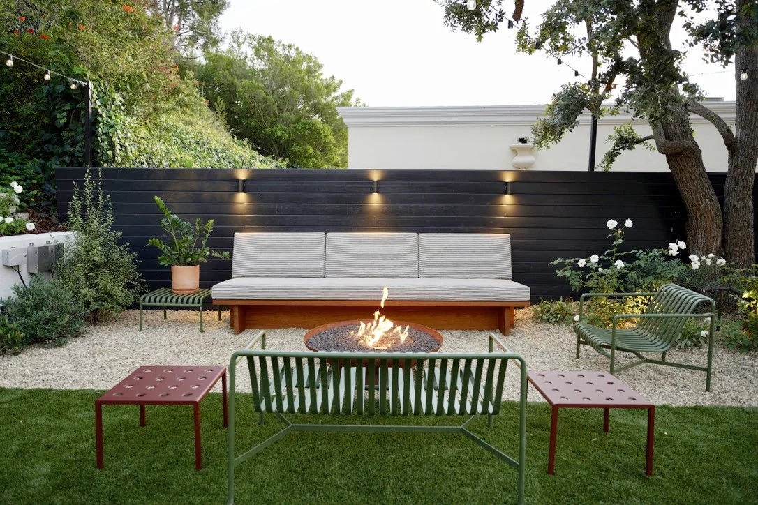 Cozy and bright yard with lush synthetic turf, fire pit lounge, play structure, horizontal slat fencing, decorative potted plantings, layered perimeter landscaping, Mexican pebbles.