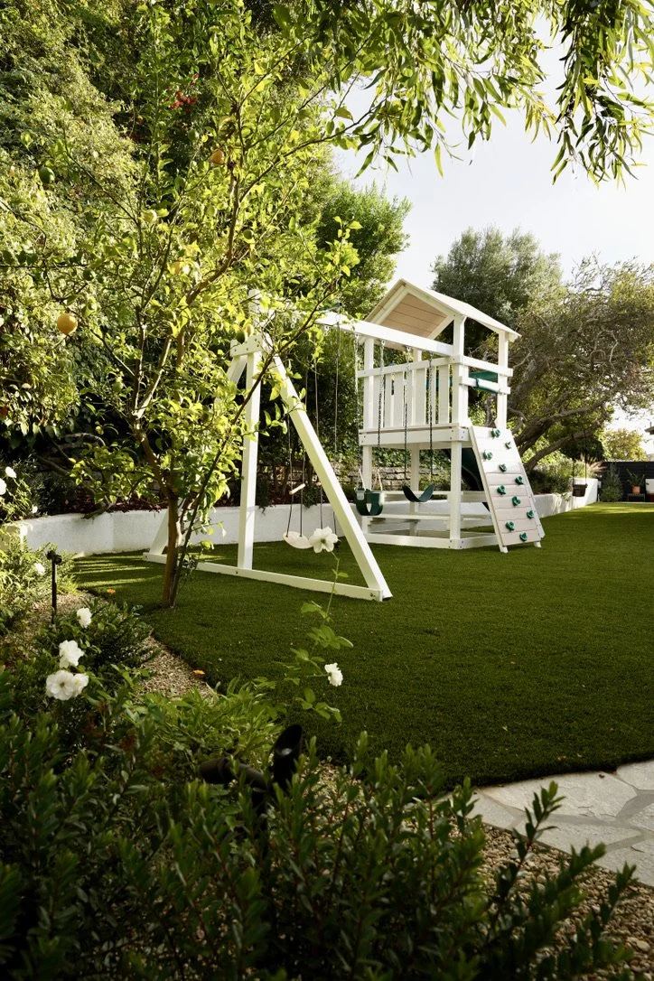Cozy and bright yard with lush synthetic turf, fire pit lounge, play structure, horizontal slat fencing, decorative potted plantings, layered perimeter landscaping, Mexican pebbles.