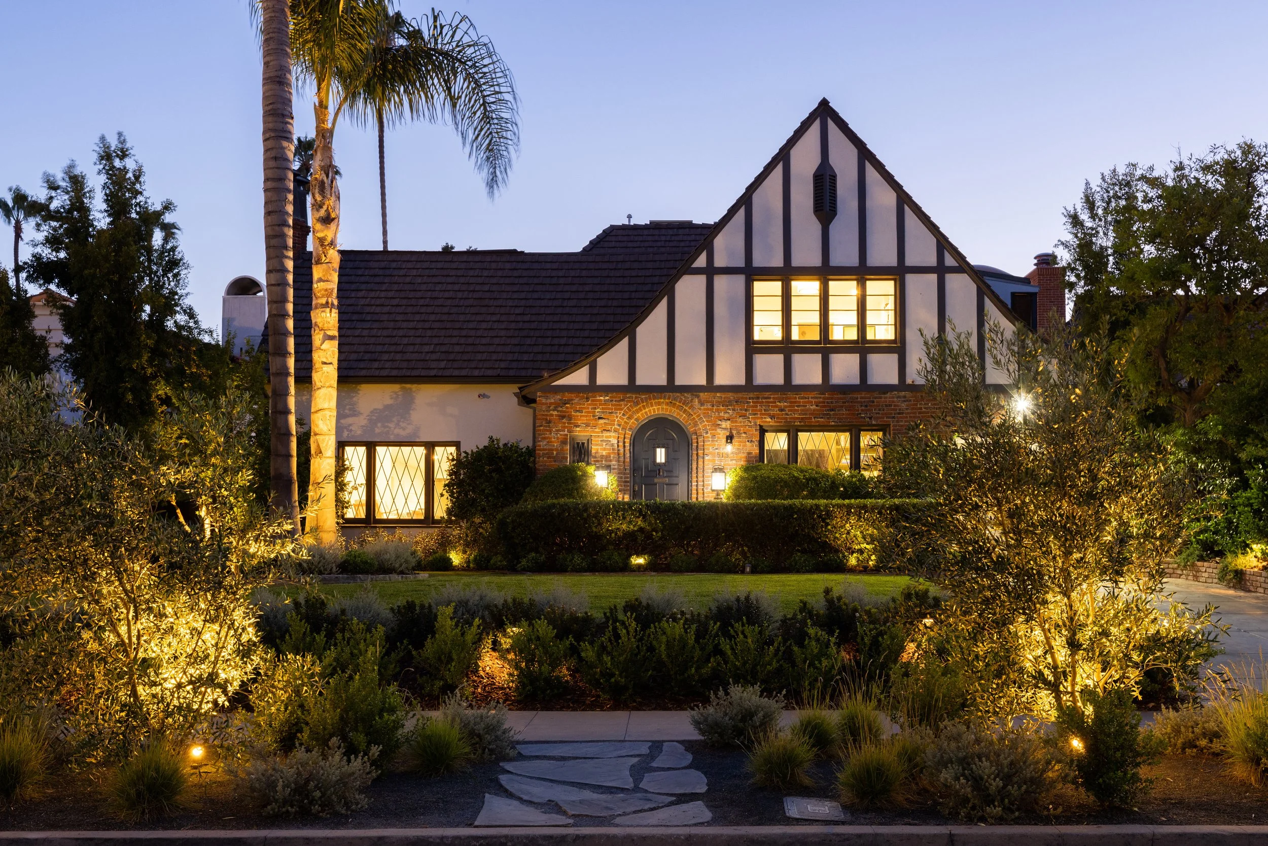 Evening photo of a tudor-style home with layered landscaping, flagstone pathway and beautiful accent landscape lighting.