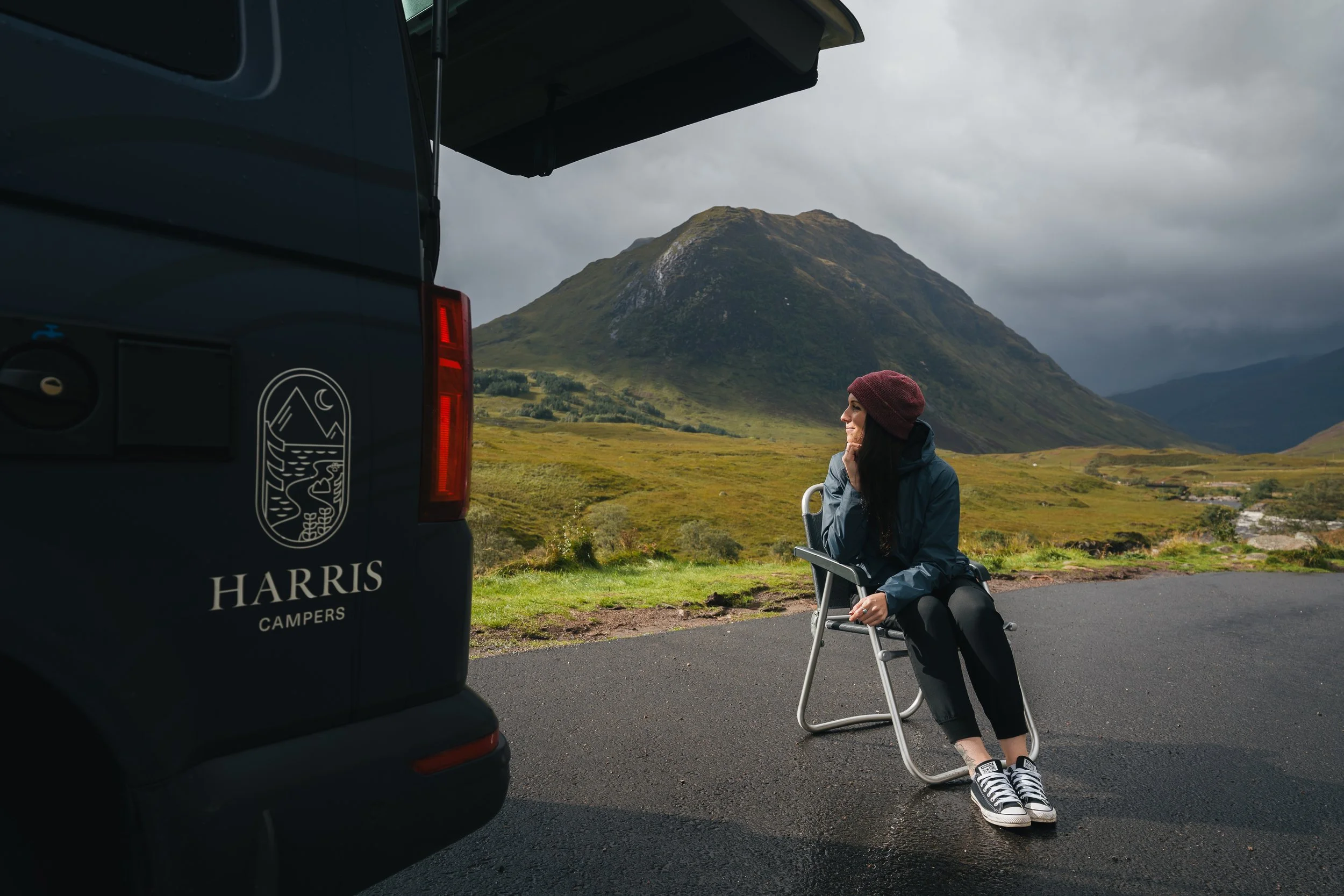 Girl sitting at Glen Coe, outside a campervan hired from Glasgow. The campervan in Scotland has the Harris Campers logo and text on it. Campervan hire Scotland