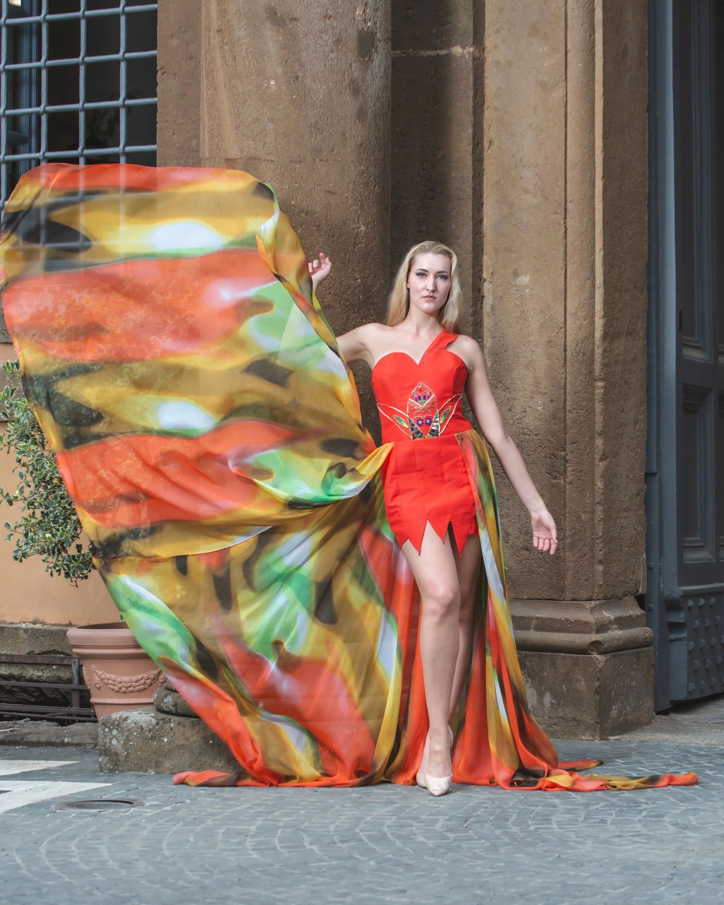 abito mono spalla realizzato in shantung rosso. Sul busto si notano ricami realizzati a mano con perline colorate, strass, filo e soutage dorata. Gonna composta da pannelli di stoffa cuciti insieme con orli a punta (con pendenti a goccia). In vita è 