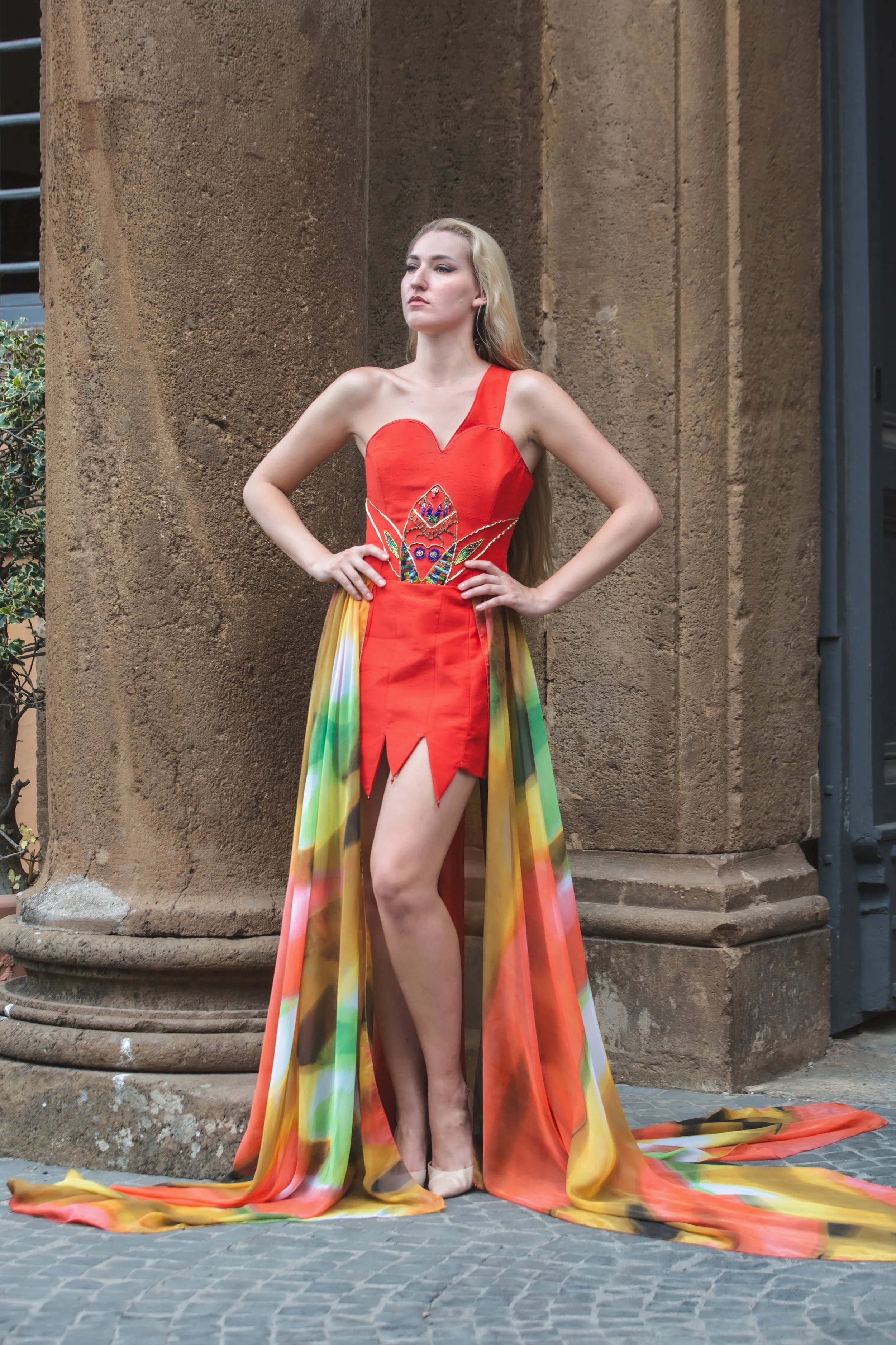 abito mono spalla realizzato in shantung rosso. Sul busto si notano ricami realizzati a mano con perline colorate, strass, filo e soutage dorata. Gonna composta da pannelli di stoffa cuciti insieme con orli a punta (con pendenti a goccia). In vita è 