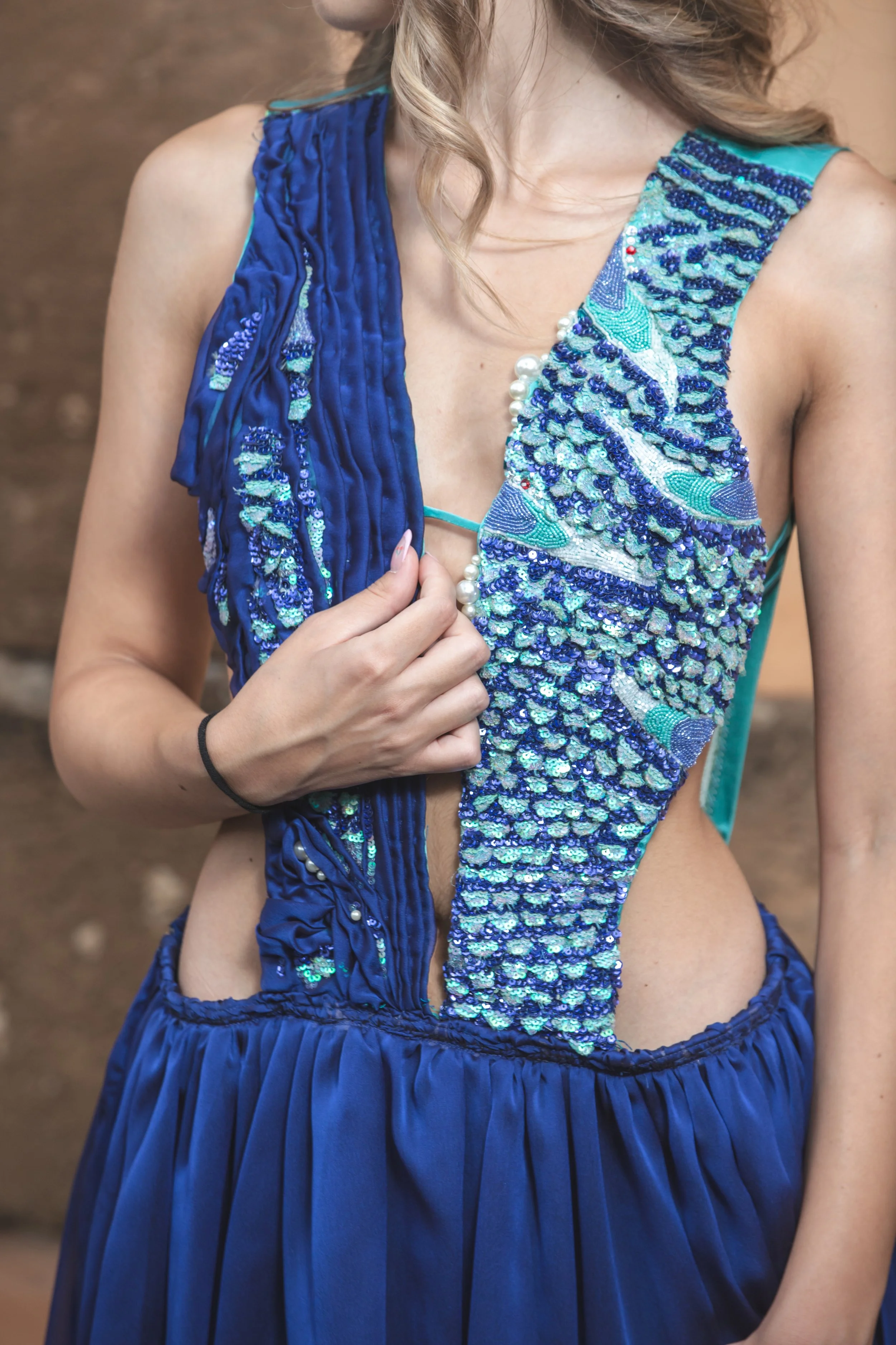 abito molto lavorato, composto da un corpino realizzato con base in duchesse celeste ricoperta da una parte da un drappeggio realizzato a mano in georgette blu, da cui fuoriescono squame realizzate e ricamate a mano con paillettes celesti e viola. L’