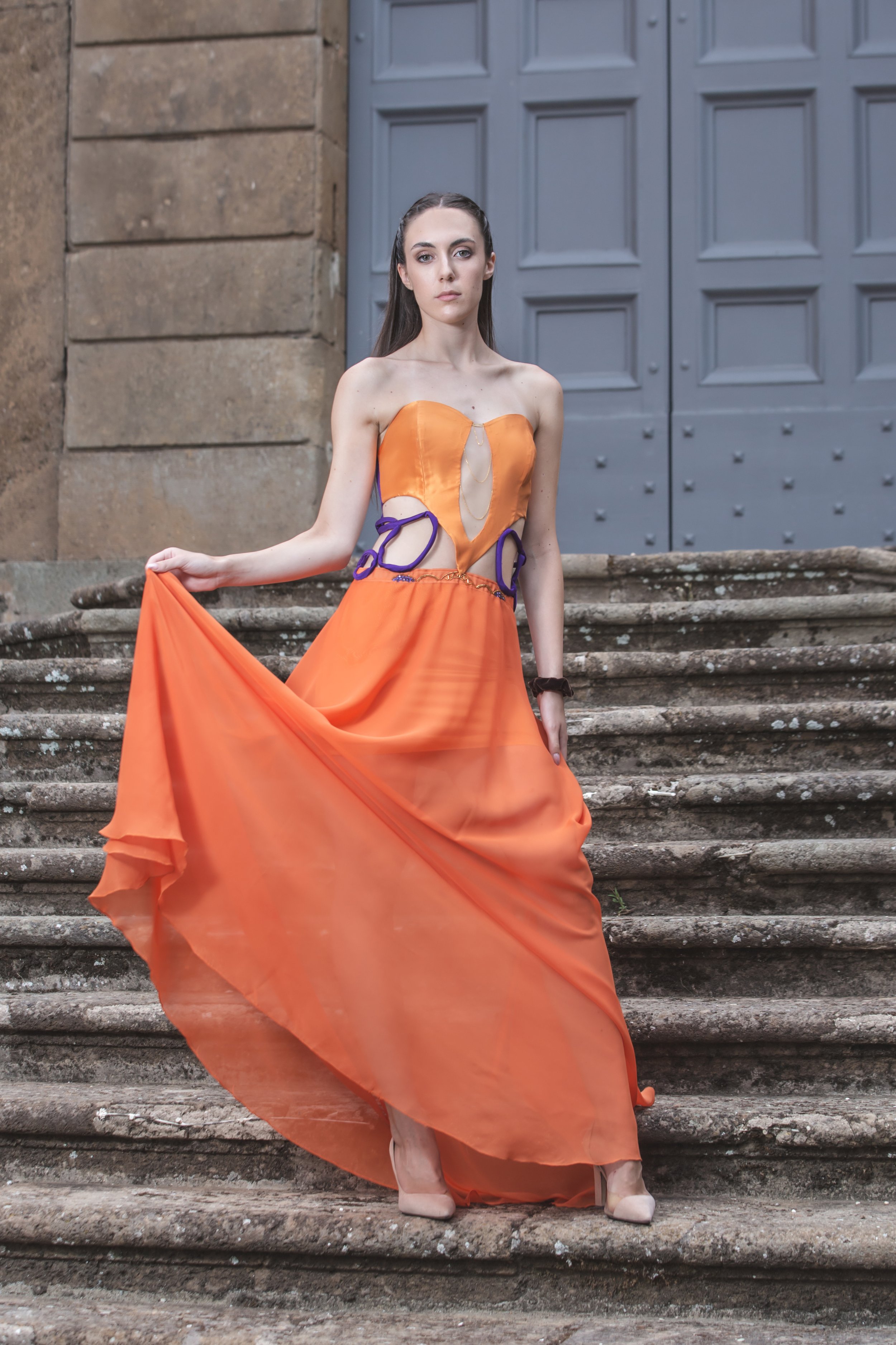 abito formato da un corpino in duchesse arancio con scollo a cuore e scollatura trasparente sul centro davanti, decorata con catenine pendenti dorate. I fianchi sono scoperti, decorati solamente da budellini viola incrociati. 
La gonna è realizzata i