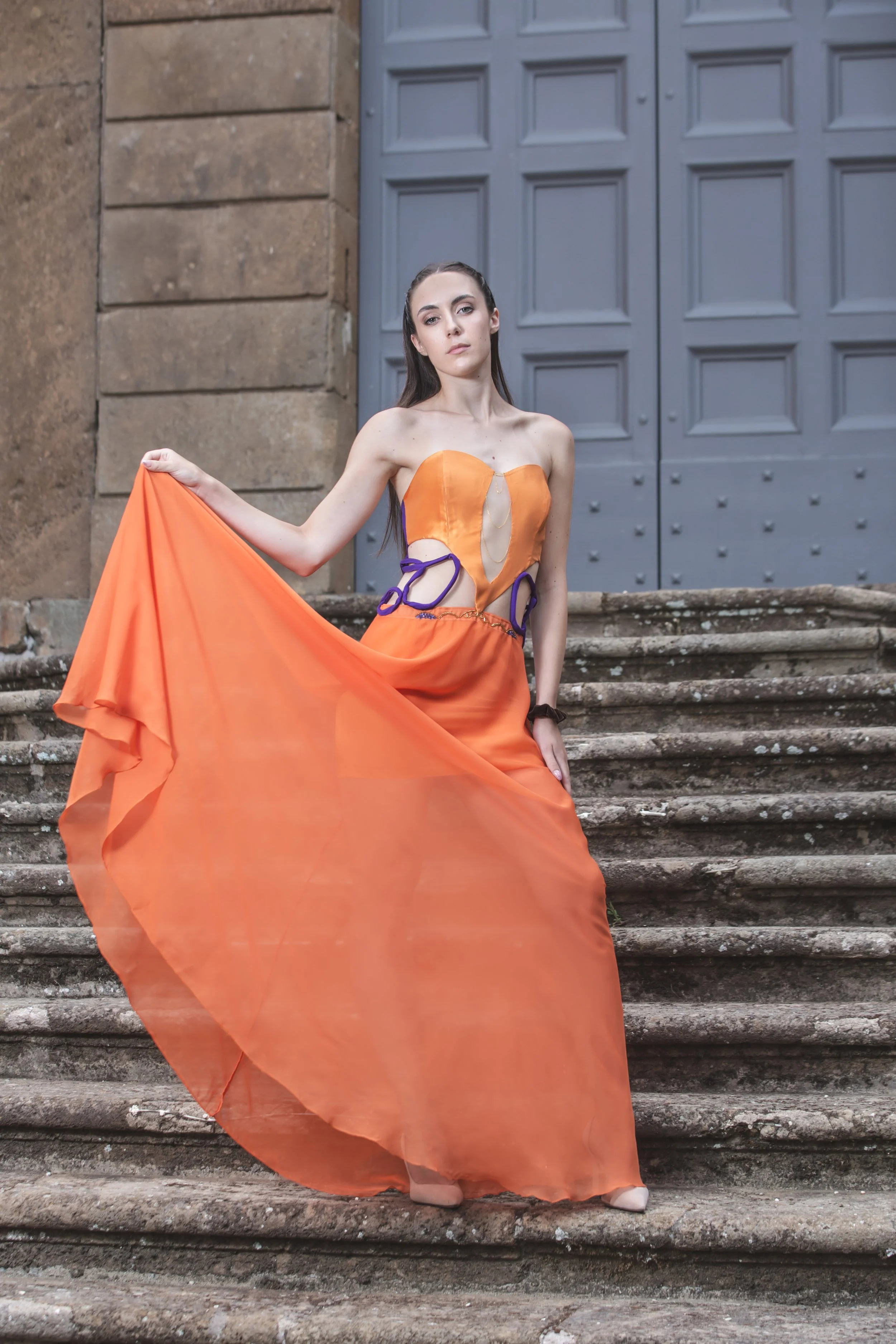 abito formato da un corpino in duchesse arancio con scollo a cuore e scollatura trasparente sul centro davanti, decorata con catenine pendenti dorate. I fianchi sono scoperti, decorati solamente da budellini viola incrociati. 
La gonna è realizzata i