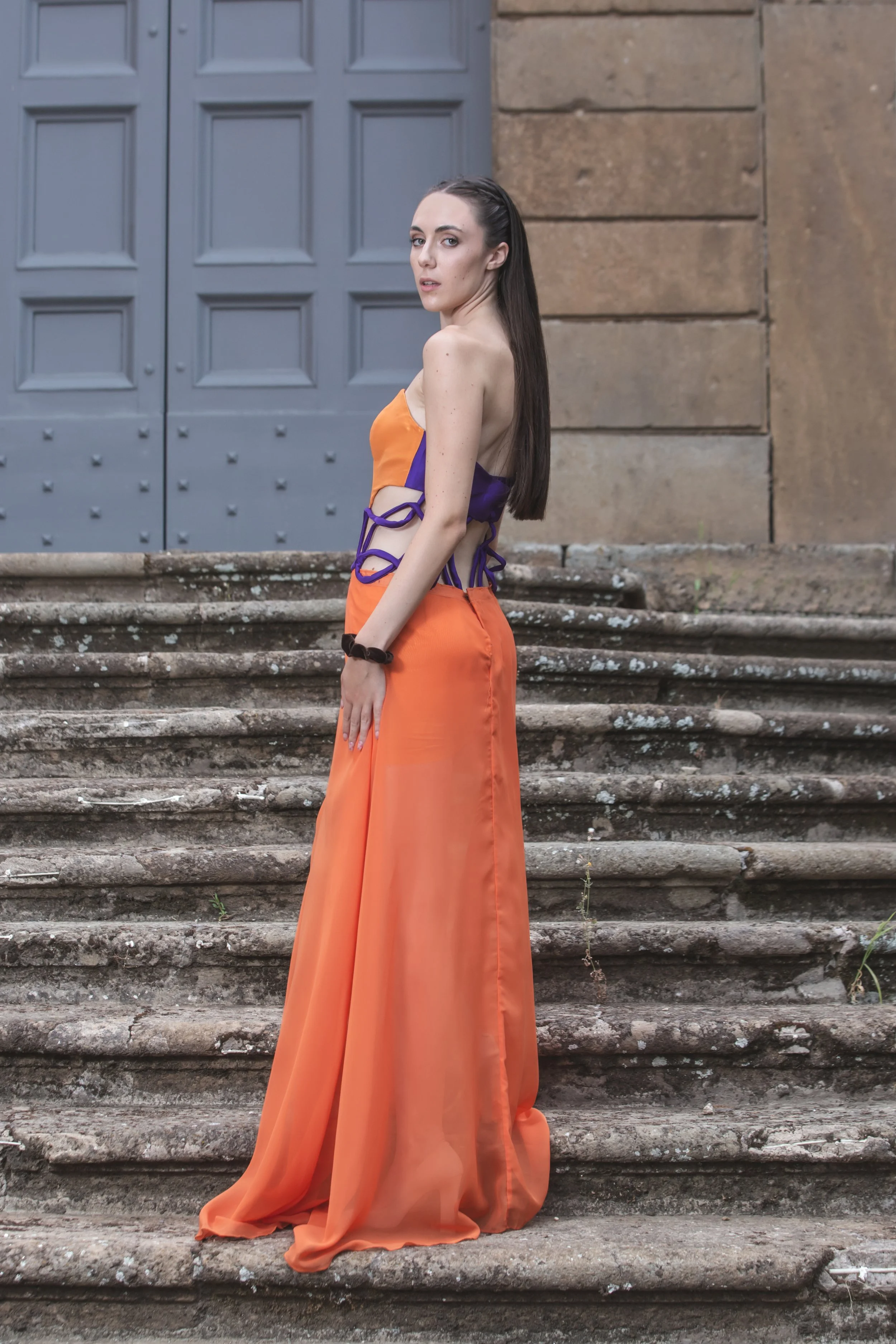 abito formato da un corpino in duchesse arancio con scollo a cuore e scollatura trasparente sul centro davanti, decorata con catenine pendenti dorate. I fianchi sono scoperti, decorati solamente da budellini viola incrociati. 
La gonna è realizzata i