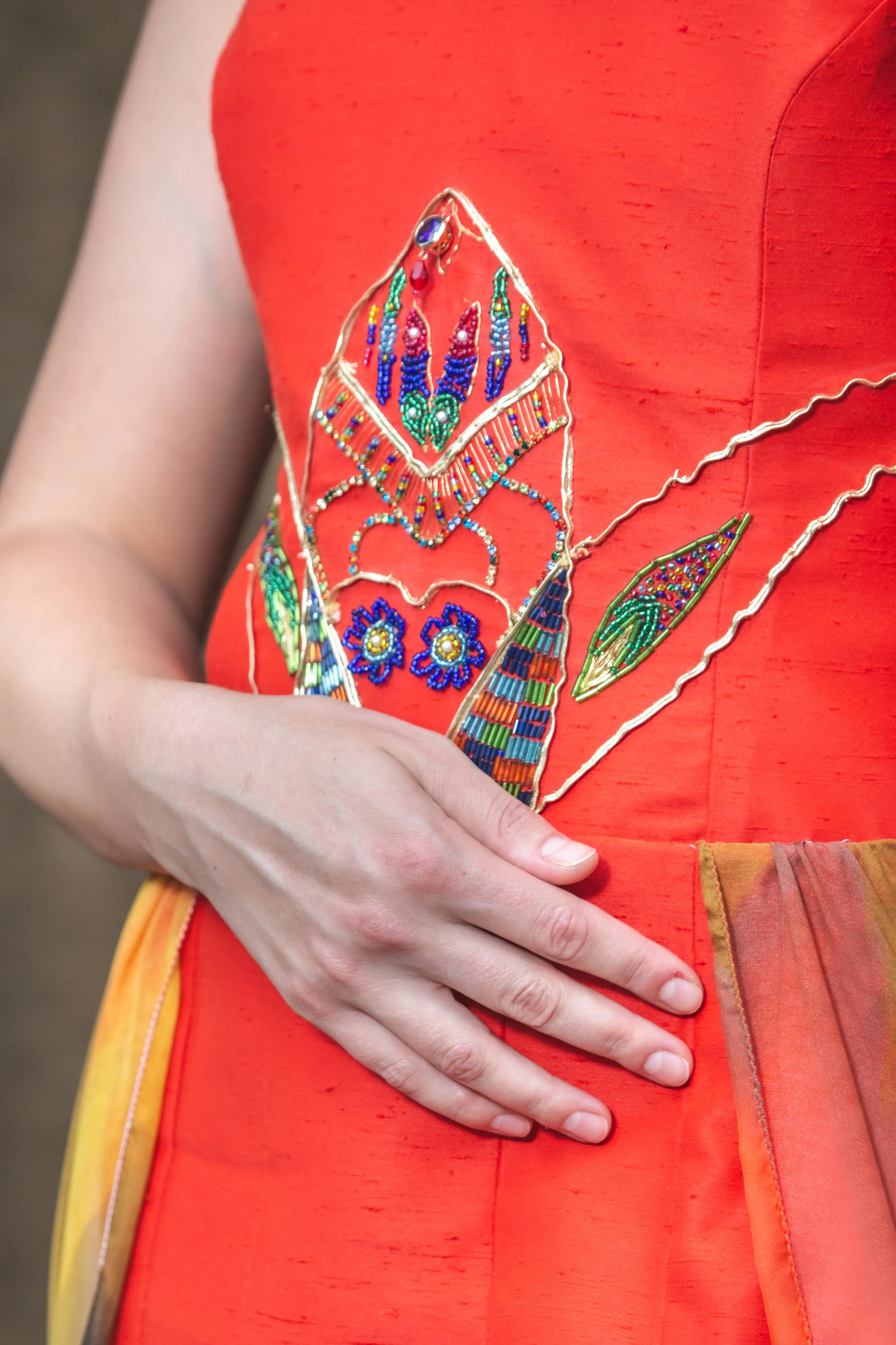 abito mono spalla realizzato in shantung rosso. Sul busto si notano ricami realizzati a mano con perline colorate, strass, filo e soutage dorata. Gonna composta da pannelli di stoffa cuciti insieme con orli a punta (con pendenti a goccia). In vita è 