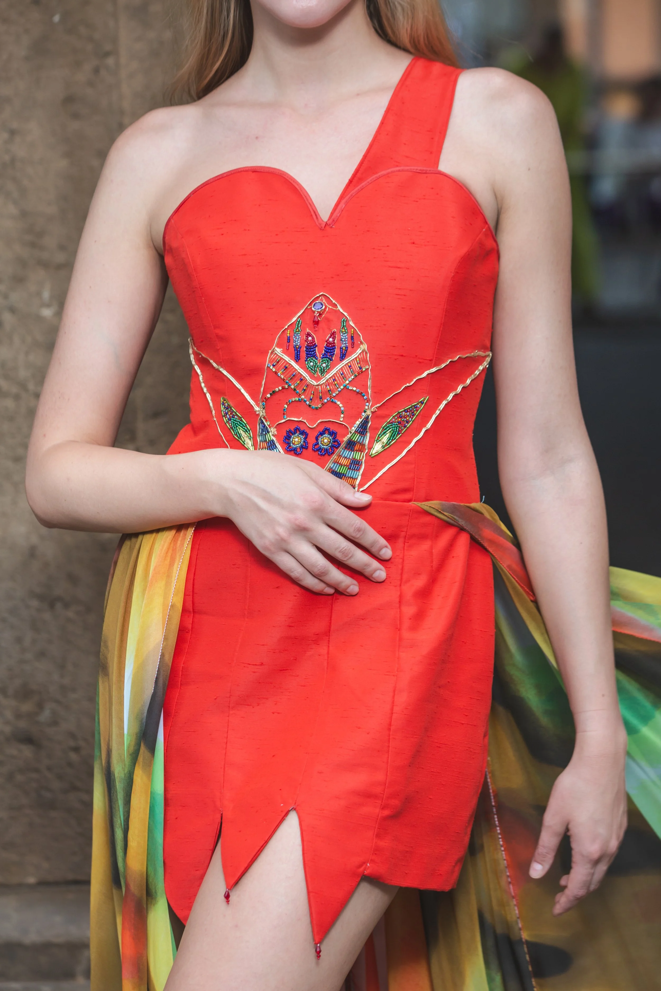 abito mono spalla realizzato in shantung rosso. Sul busto si notano ricami realizzati a mano con perline colorate, strass, filo e soutage dorata. Gonna composta da pannelli di stoffa cuciti insieme con orli a punta (con pendenti a goccia). In vita è 