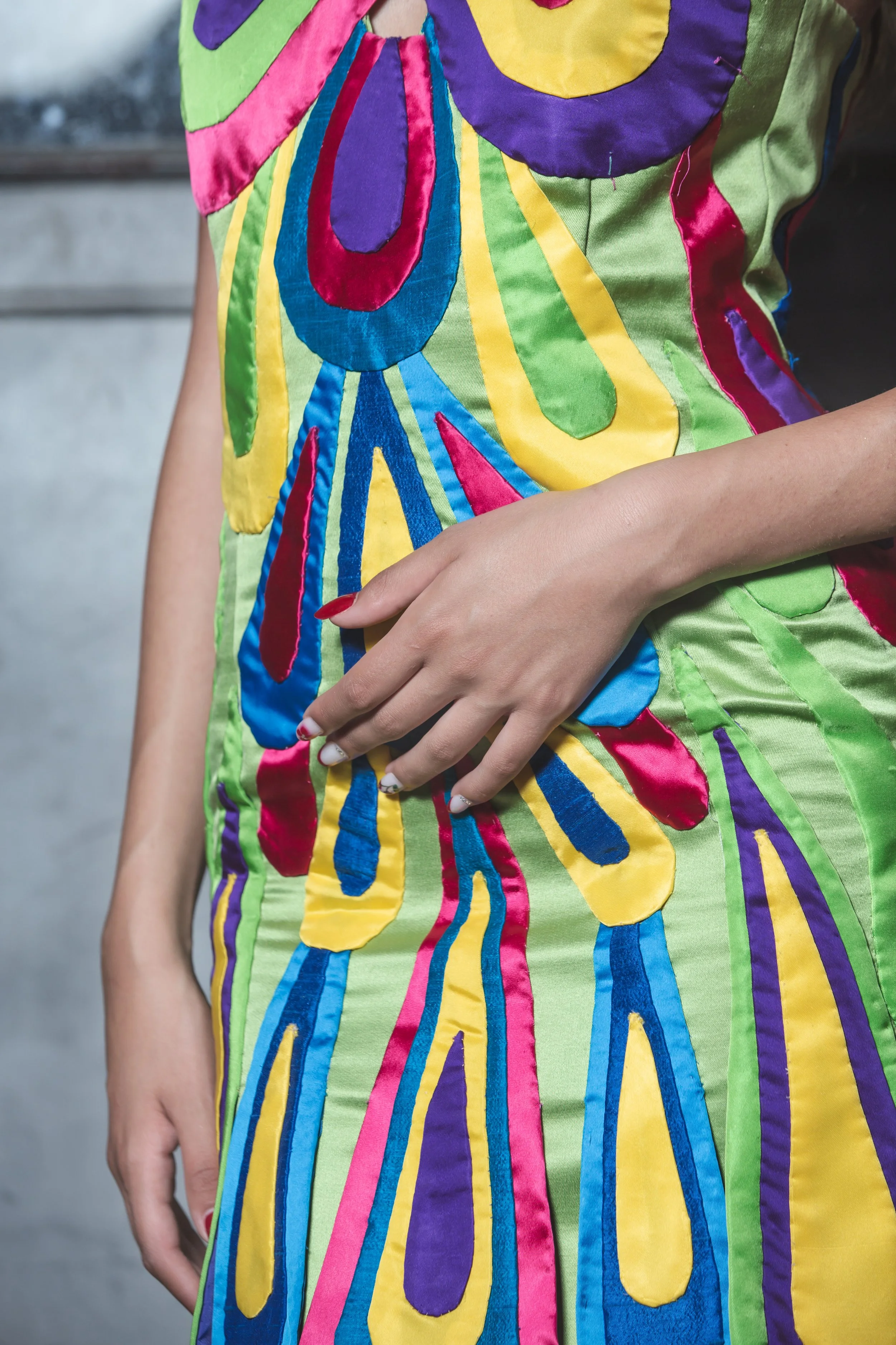 La base del tubino è realizzata in raso verde chiaro con scollo all’americana incrociato sul davanti tramite due grandi gocce realizzate a mano.
Ogni goccia è realizzata e cucita a mano con tessuti diversi: raso fucsia e celeste, shantung nei toni de