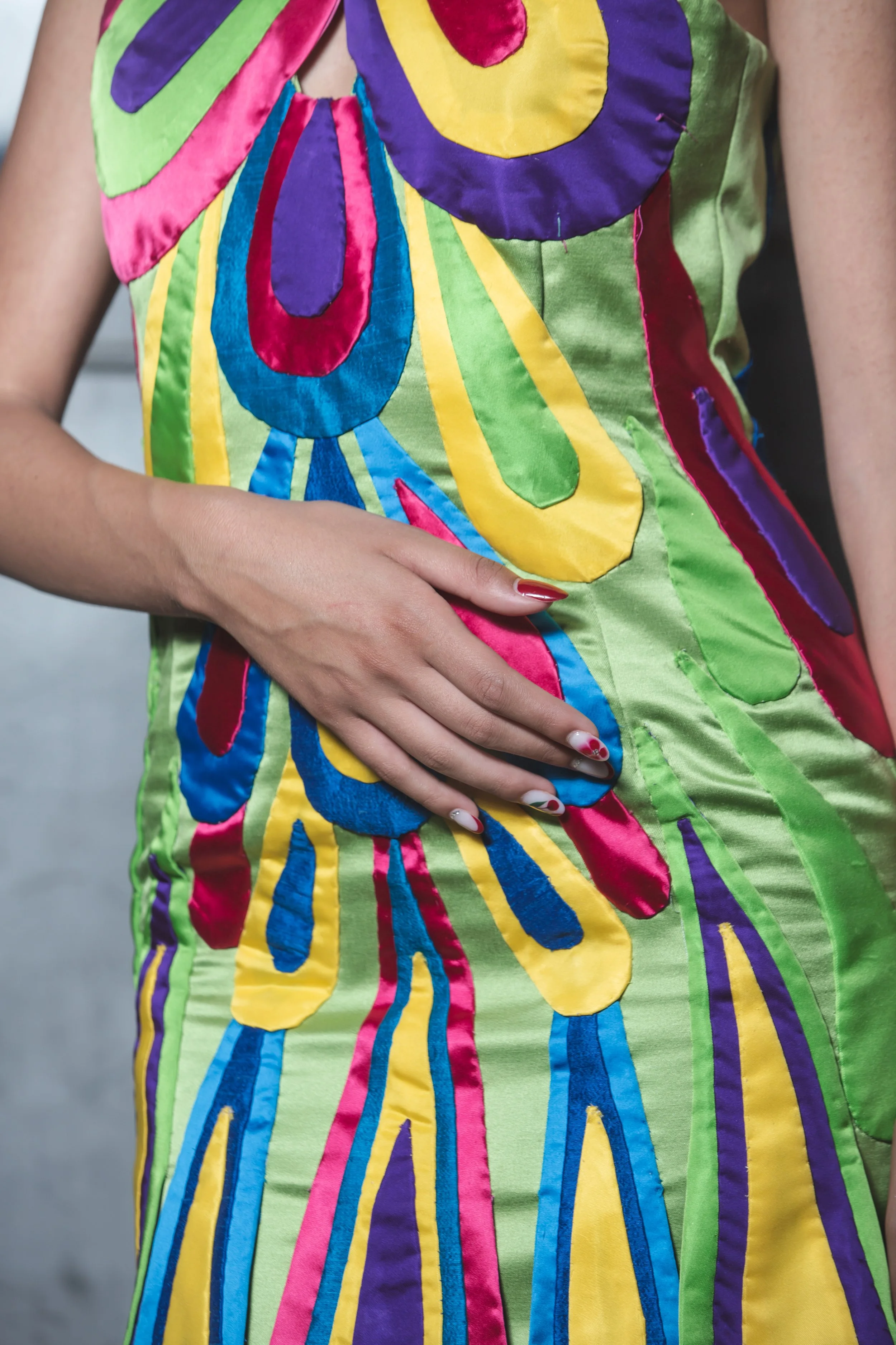 La base del tubino è realizzata in raso verde chiaro con scollo all’americana incrociato sul davanti tramite due grandi gocce realizzate a mano.
Ogni goccia è realizzata e cucita a mano con tessuti diversi: raso fucsia e celeste, shantung nei toni de