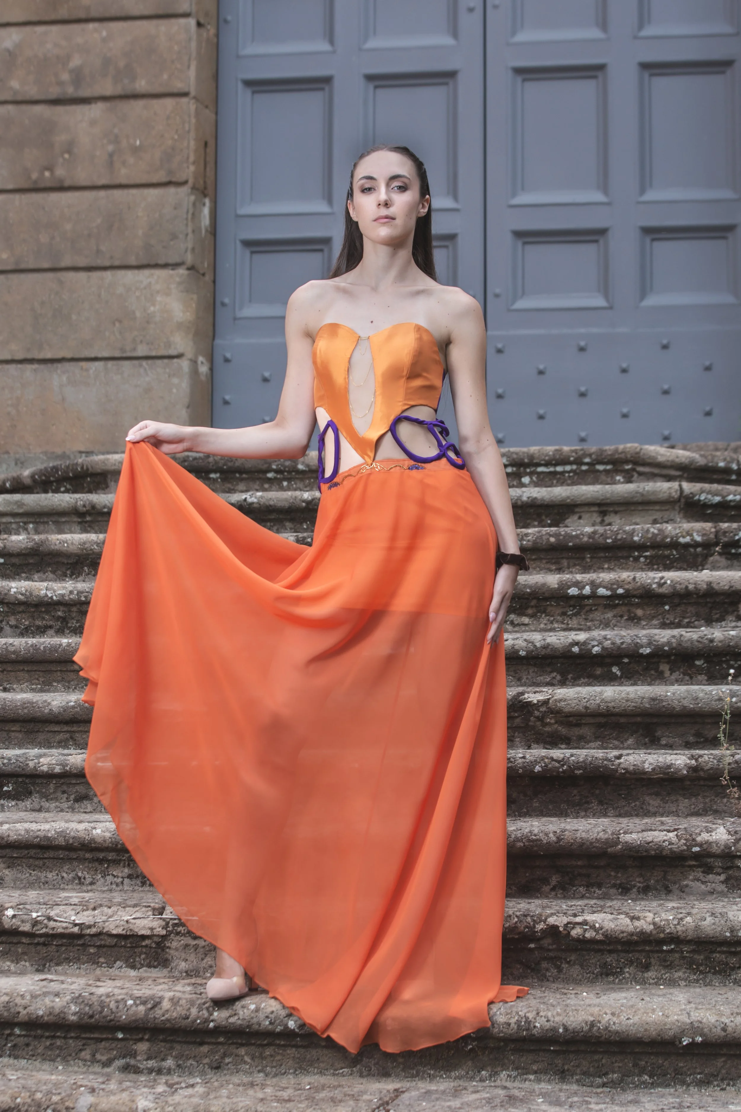 abito formato da un corpino in duchesse arancio con scollo a cuore e scollatura trasparente sul centro davanti, decorata con catenine pendenti dorate. I fianchi sono scoperti, decorati solamente da budellini viola incrociati. 
La gonna è realizzata i
