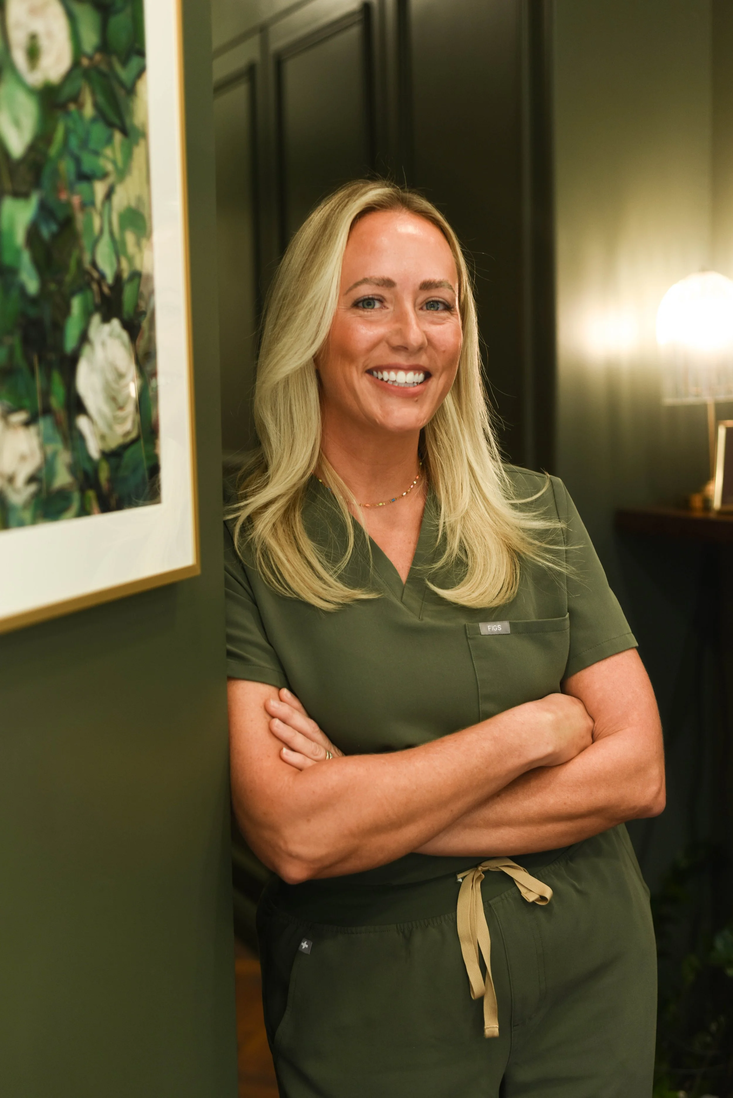 A smiling woman wearing green scrubs standing in a room with dark-colored walls, crossed arms, and a framed floral artwork on the wall.