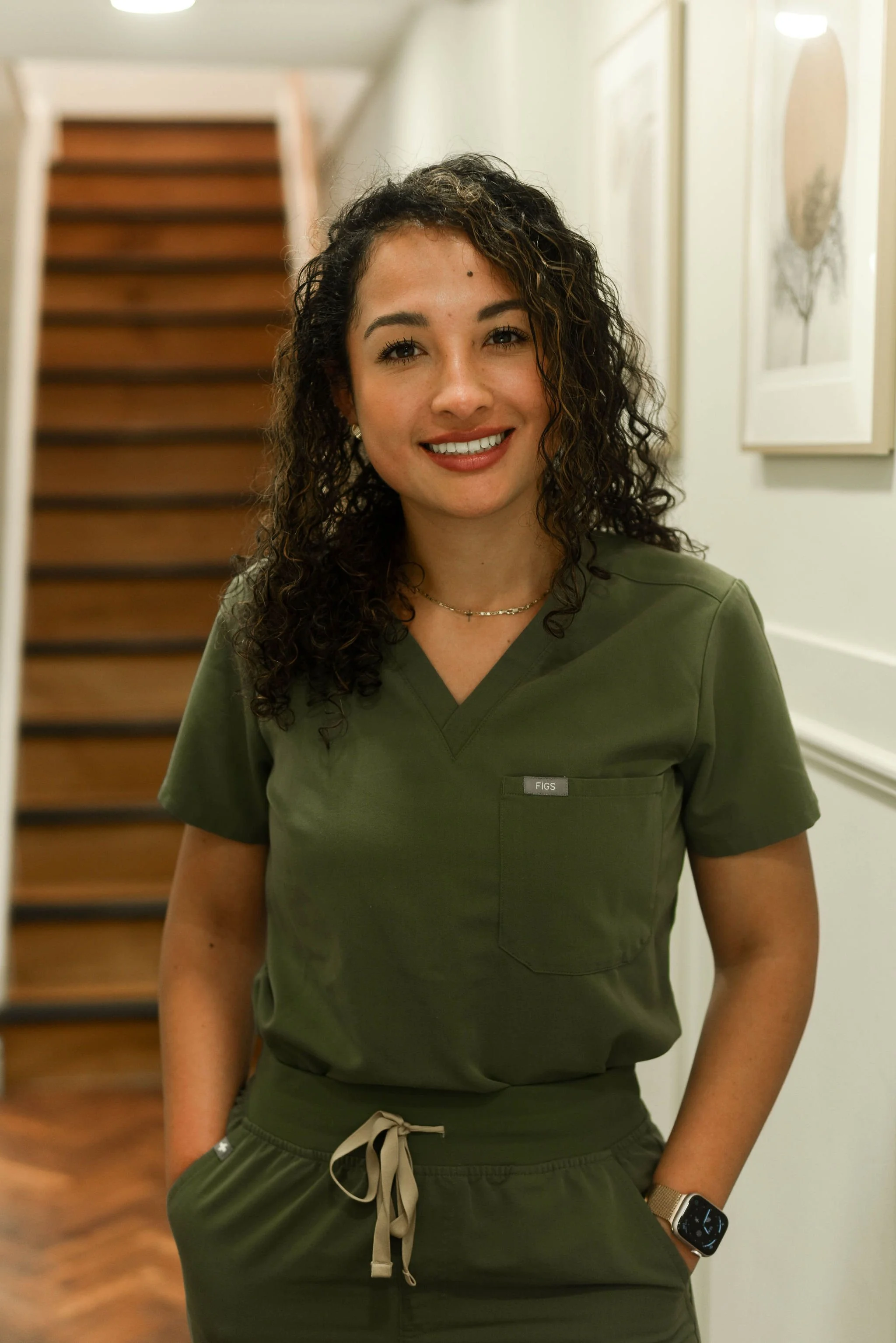 A woman with curly dark hair wearing green medical scrubs, standing in a hallway with stairs and framed pictures on the wall.